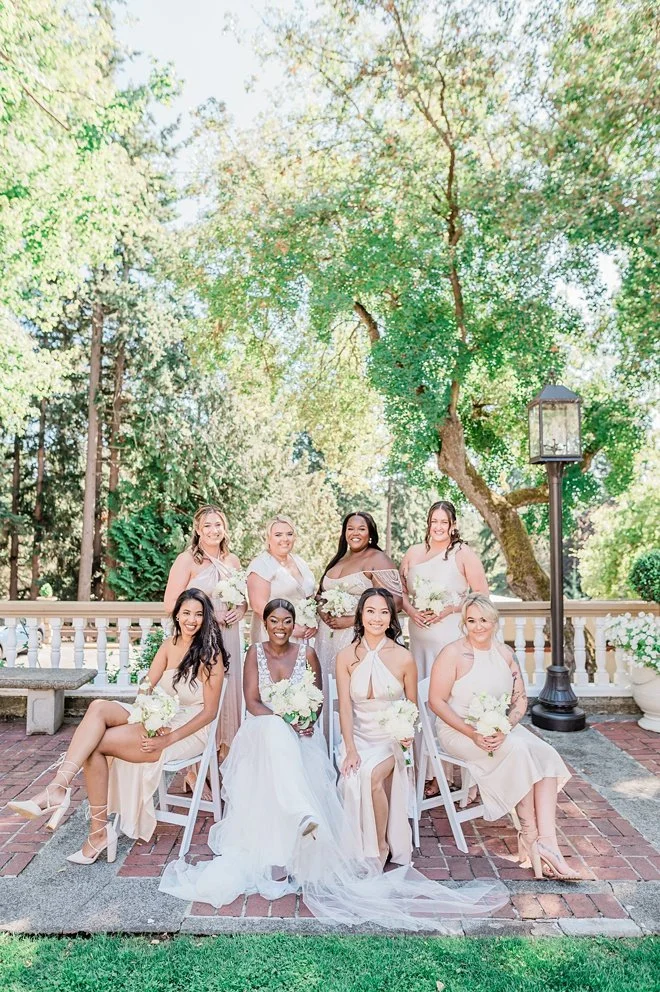 A group of nine women in light-colored dresses, some in white and others in blush pink, pose outdoors for a wedding photo. They are smiling, holding bouquets, with a tree, a white railing, and a lamp post in the background.