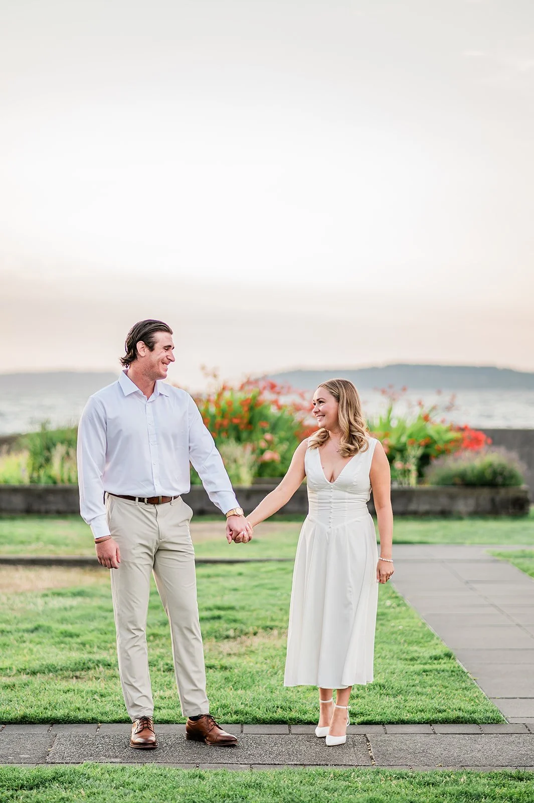 Couple holding hands on the lighthouse lawn with water and red flowers behind them