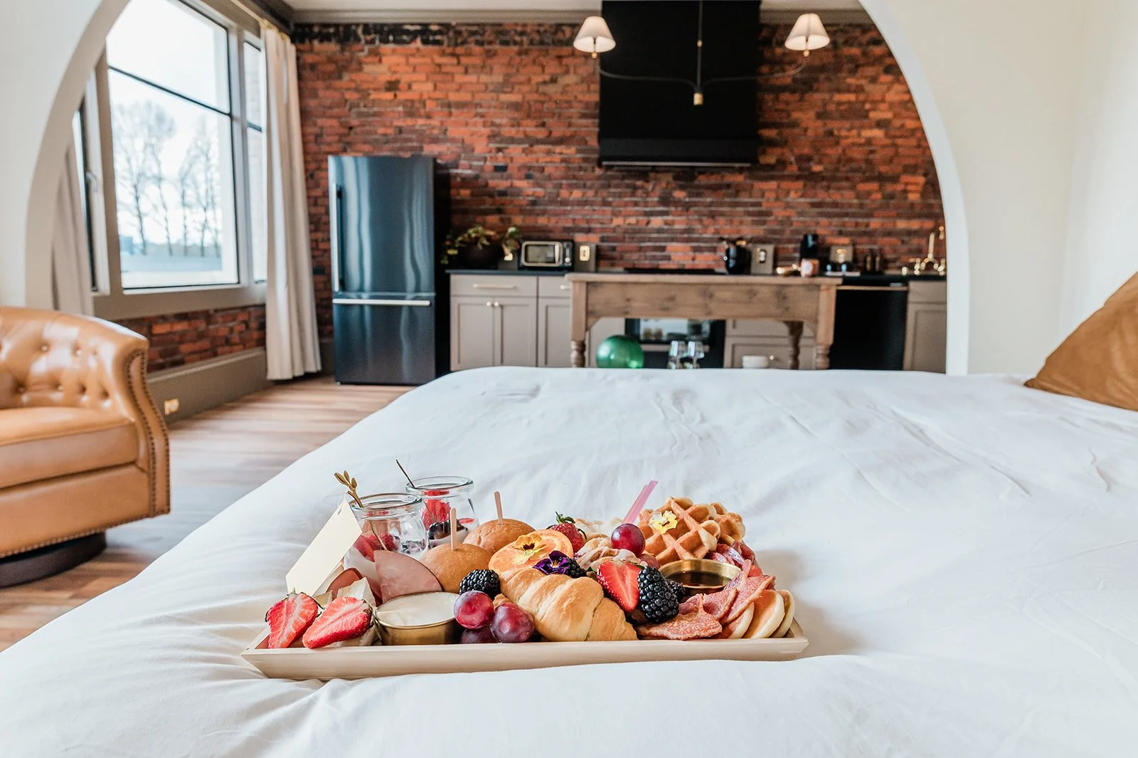 908 suite brunch tray — personal breakfast spread on white luxury bedding with original brick and arched kitchen beyond