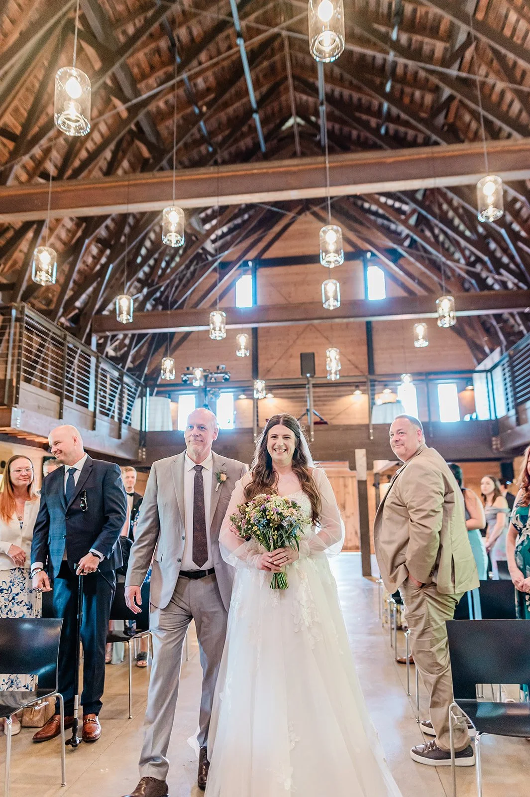 Father walking the bride down the aisle under pendant lights in the Marionfield Farm barn
