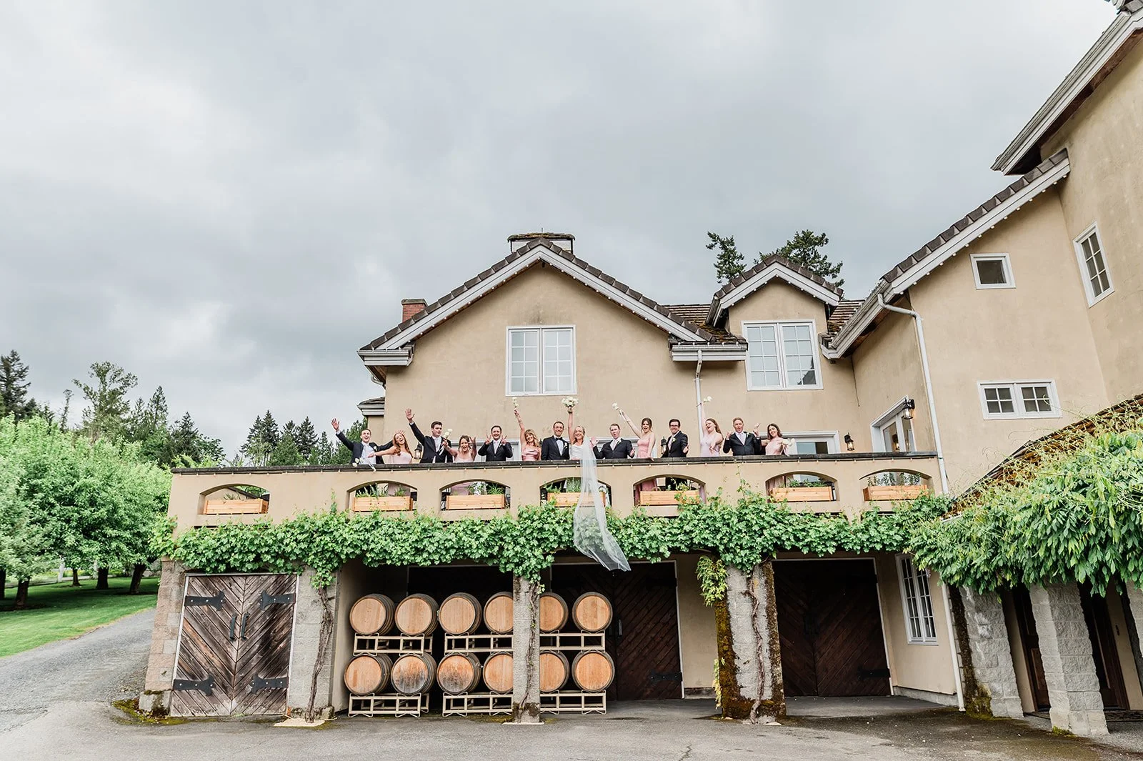 Wedding party cheering on the Chateau Lill balcony above the wine barrel wall, with the bride’s veil draped over the railing