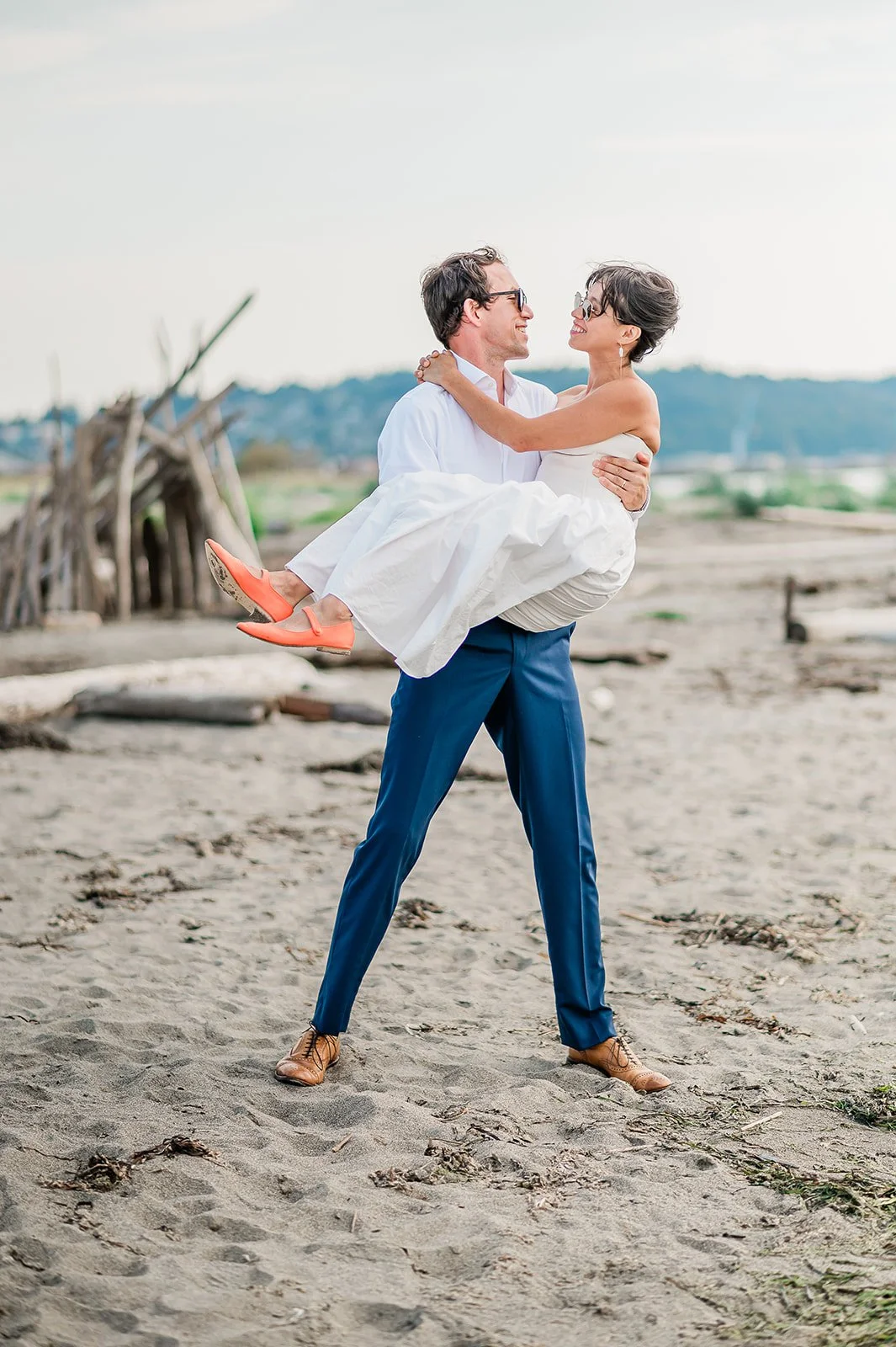 Groom lifting bride in wedding dress on Jetty Island beach with driftwood and soft summer sky Everett Washington wedding photographer
