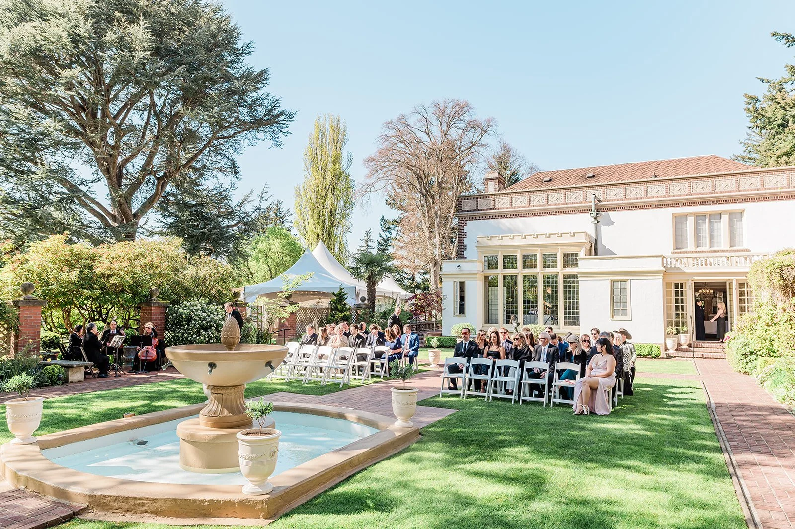 Wide shot of a wedding ceremony in the fountain garden at Lairmont Manor in Bellingham WA, with guests seated on either side of the brick aisle, the blue fountain in the foreground and the manor behind