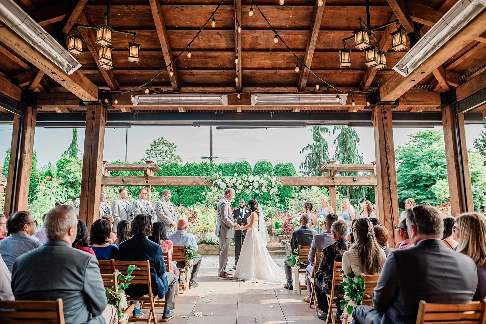 A wedding ceremony taking place under a wooden pavilion with guests seated, a bride and groom exchanging vows, and a wedding party standing nearby in front of lush green trees and floral arrangements.