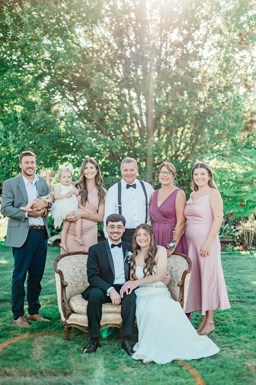 Newlyweds seated on a vintage loveseat with family standing behind them in open shade