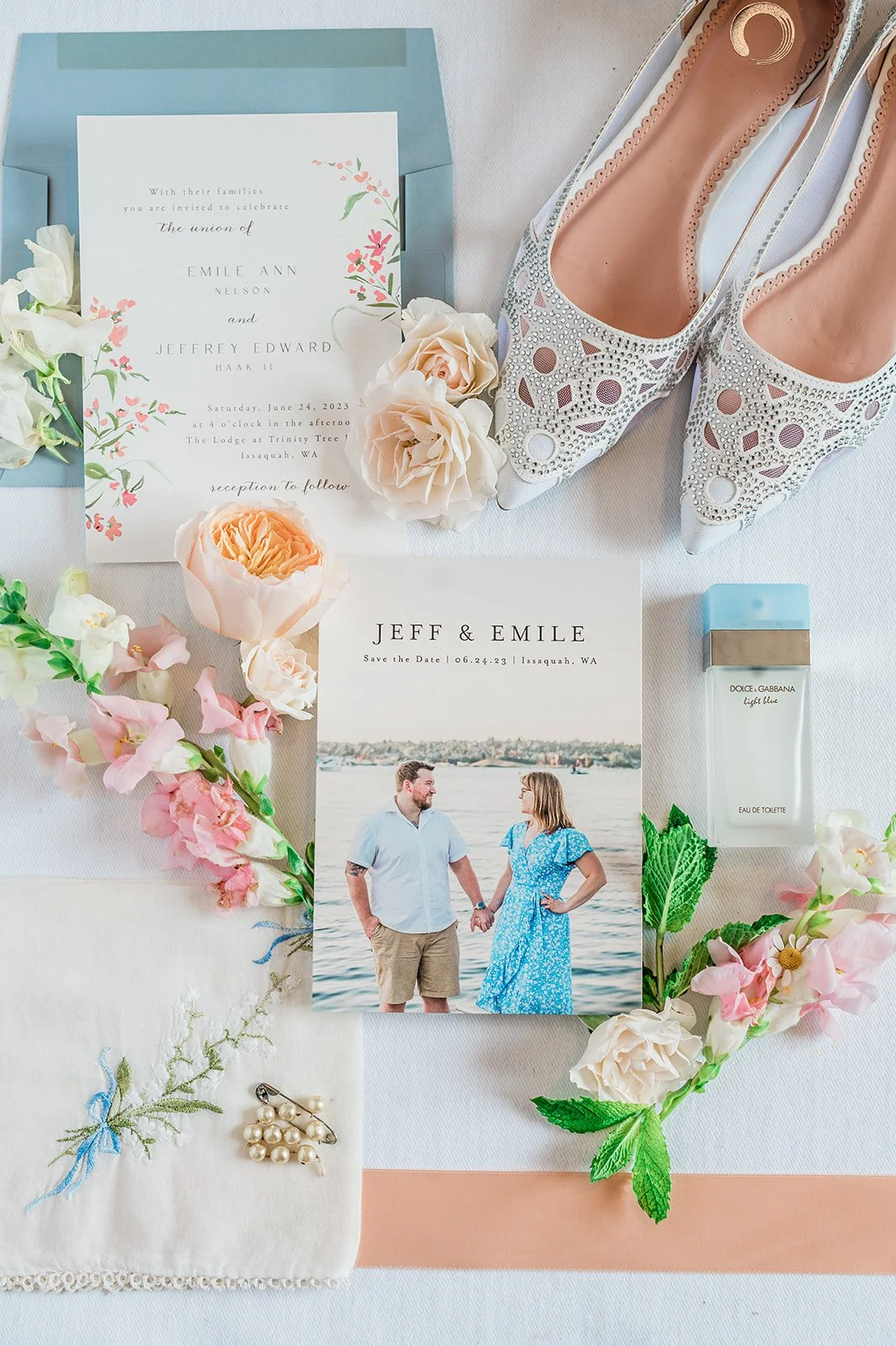 Wedding invitation, shoes, flowers, perfume, and a photo of a couple at the water, all arranged on a white surface.