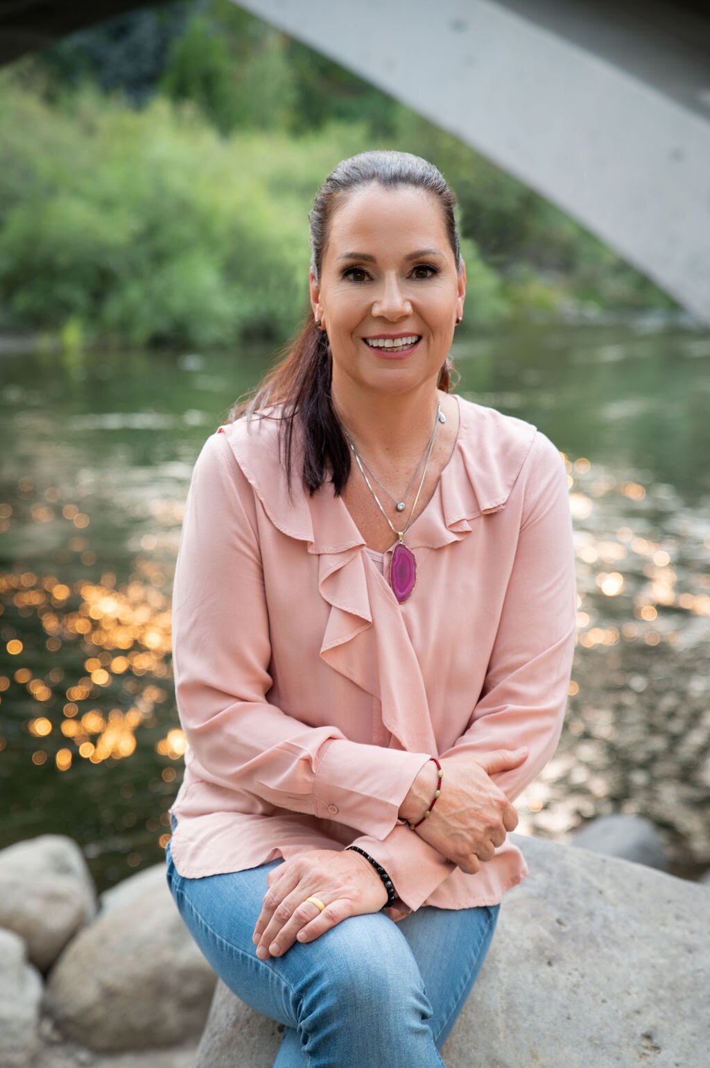 A woman sitting on a rock in a shallow river, smiling at the camera, surrounded by trees and rocks. Feng Shui Consultation, International Feng Shui Guild, Kerstin Tracy