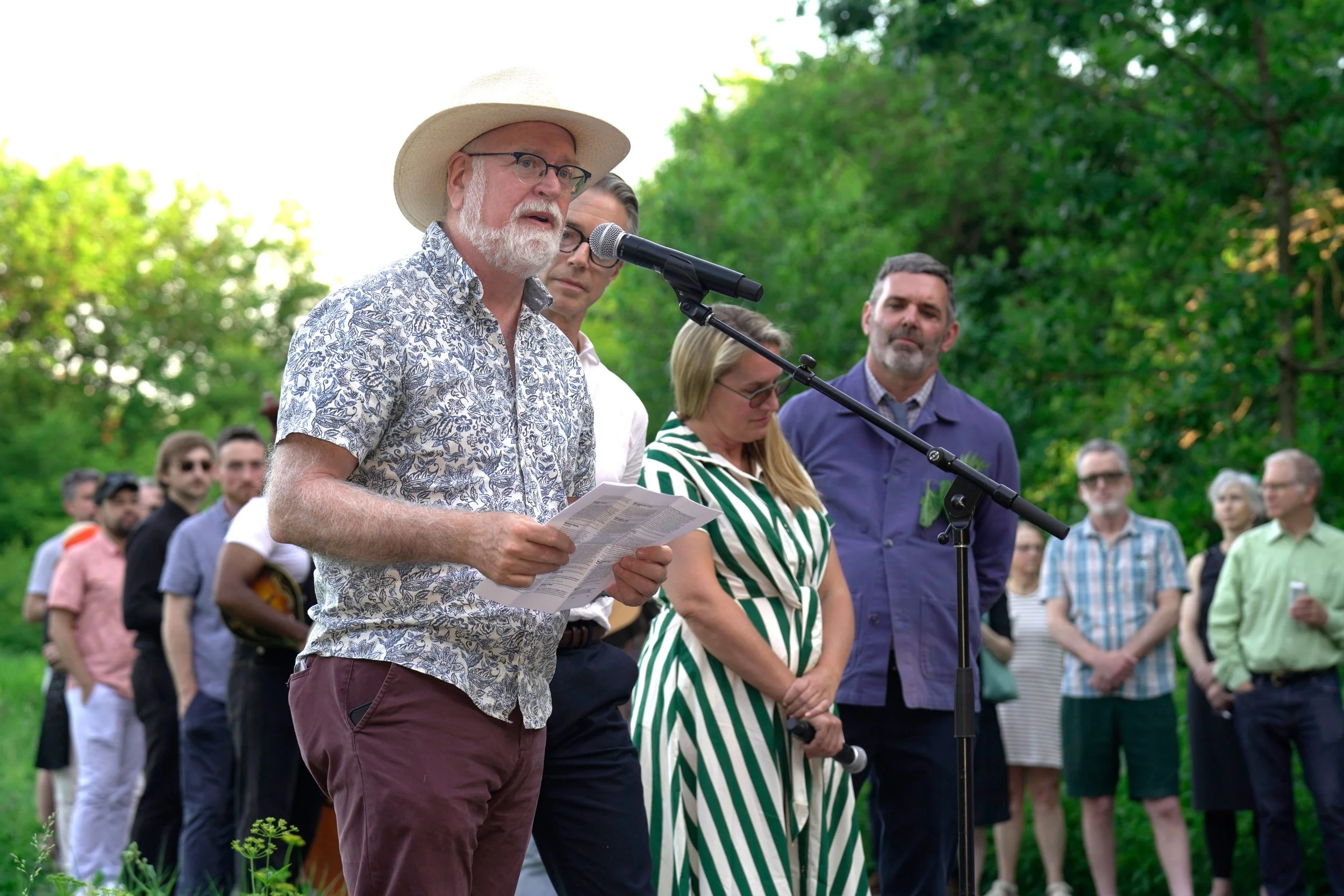A man with glasses, a beard, and a hat speaking at an outdoor event. Several people are gathered behind him, listening attentively.