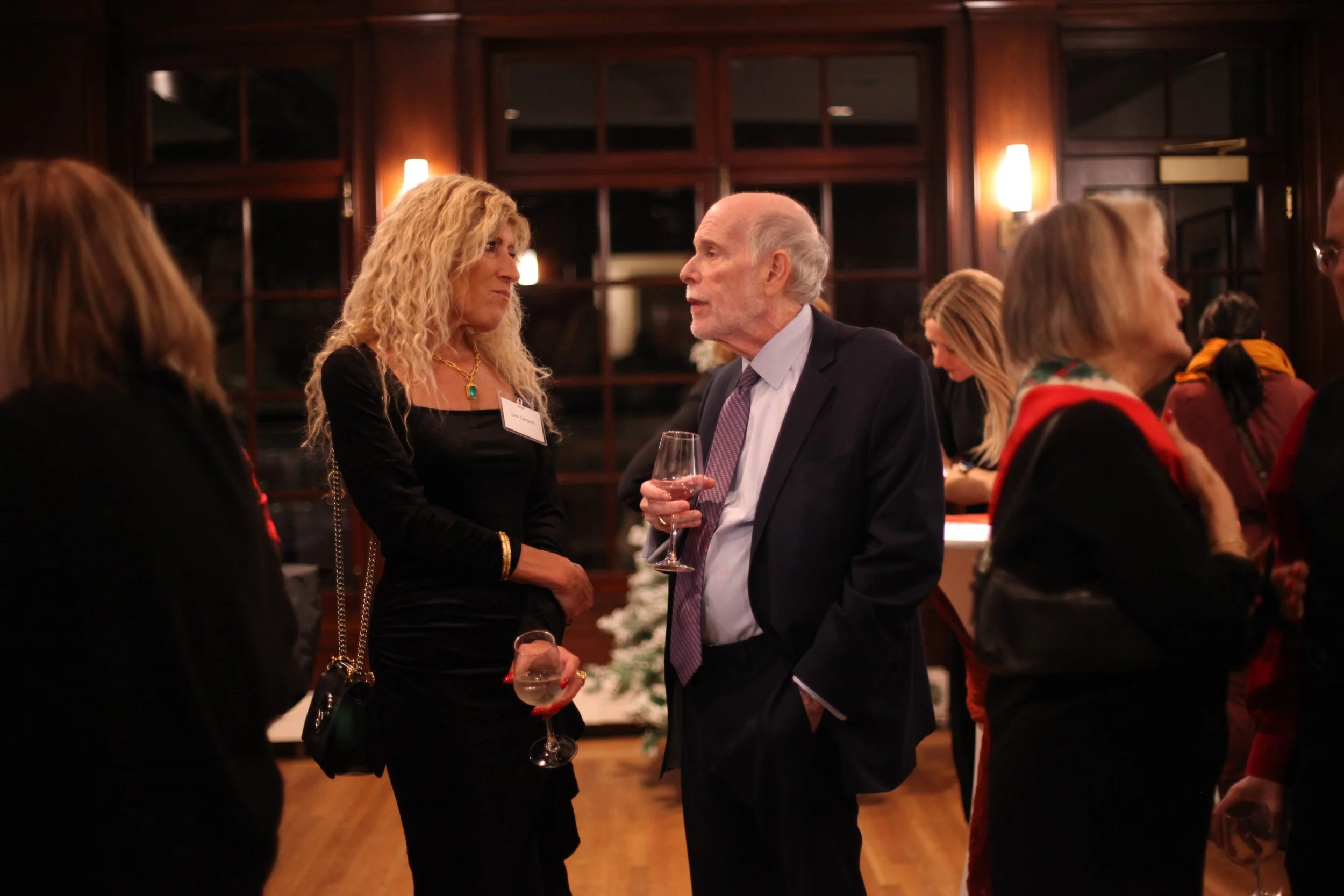 People are mingling at a formal social event or gathering, with some holding wine glasses, in a warm, wood-paneled room with dim lighting.