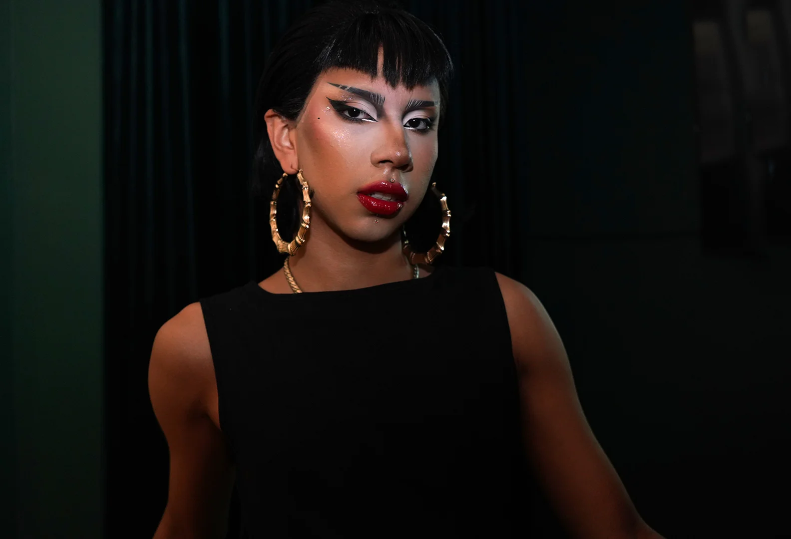 A woman with short black hair and bold makeup, wearing large gold hoop earrings and a black sleeveless top, is standing against a dark background.