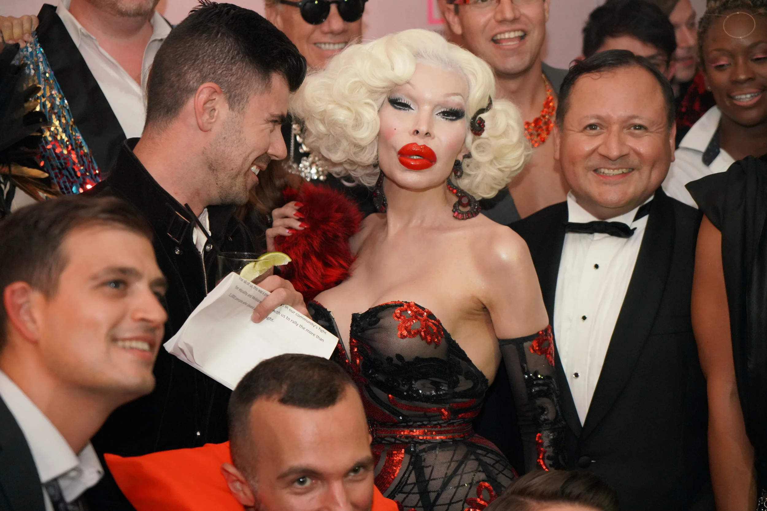 A group of people dressed in formal and glamorous clothing, smiling and celebrating at a party or event. The central figure is a woman with platinum blonde hair styled in vintage curls, wearing bold makeup and a red and black dress with lace and sequ