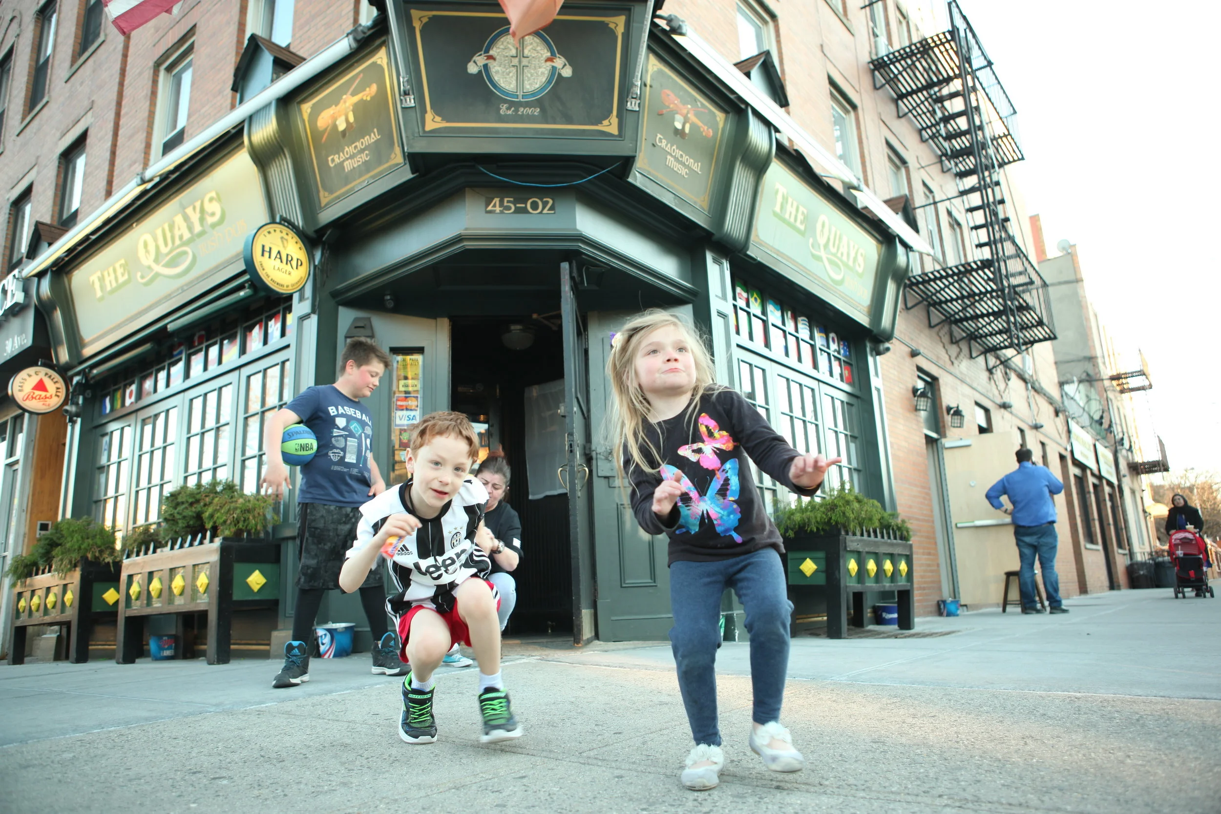 Children playing and running outside in front of a pub called The Quays, with a building and fire escape visible in the background.