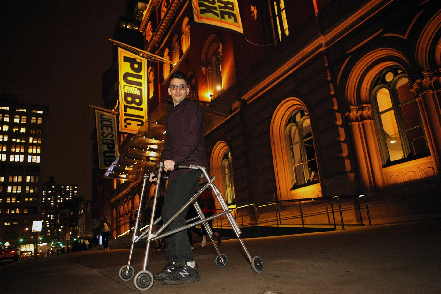A man standing outdoors at night next to a walker in front of a lit historic building with signs for 'Public Theatre' and 'Joe's Pub'.