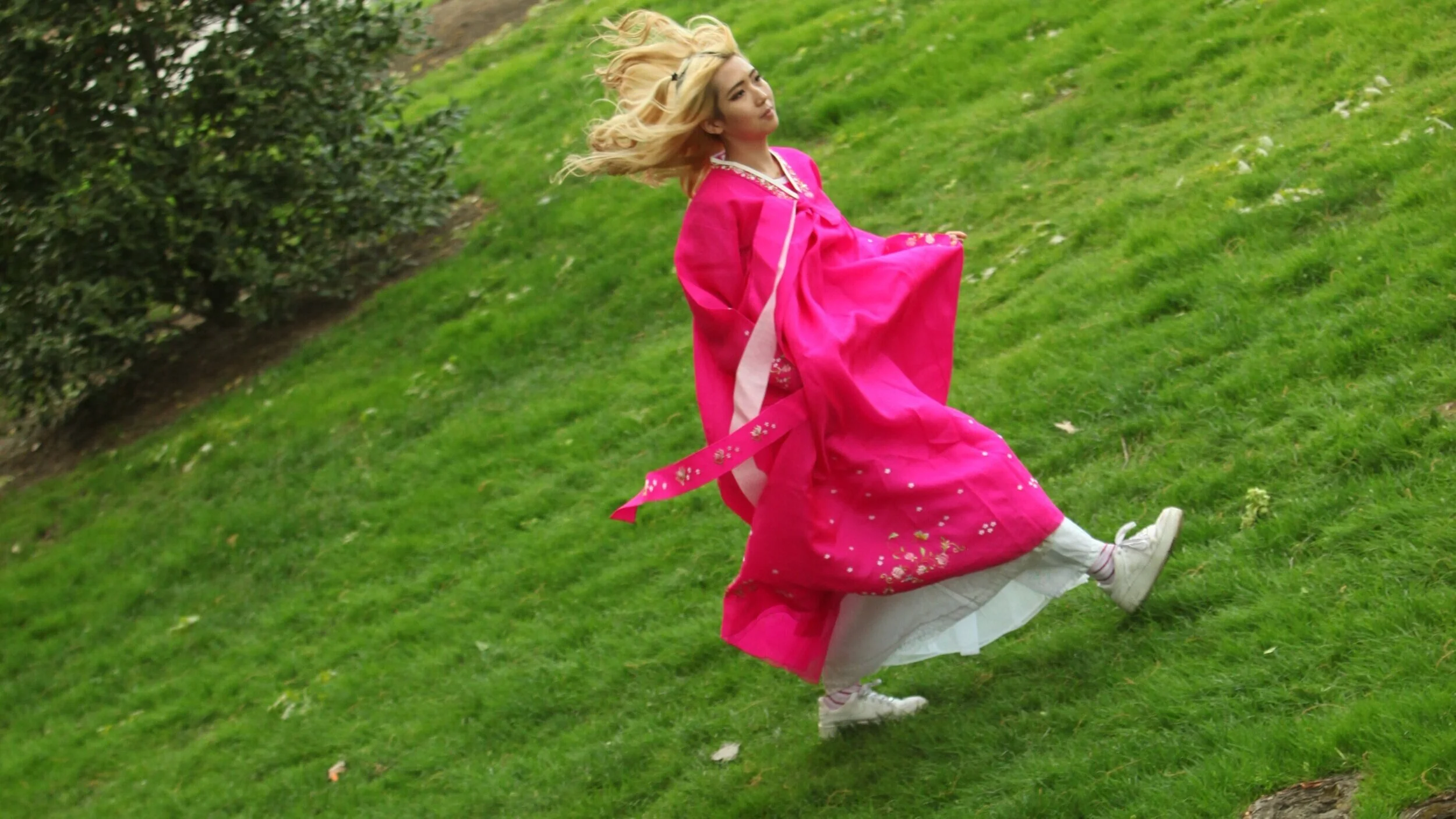 A woman in a bright pink traditional dress spinning on a grassy lawn area.