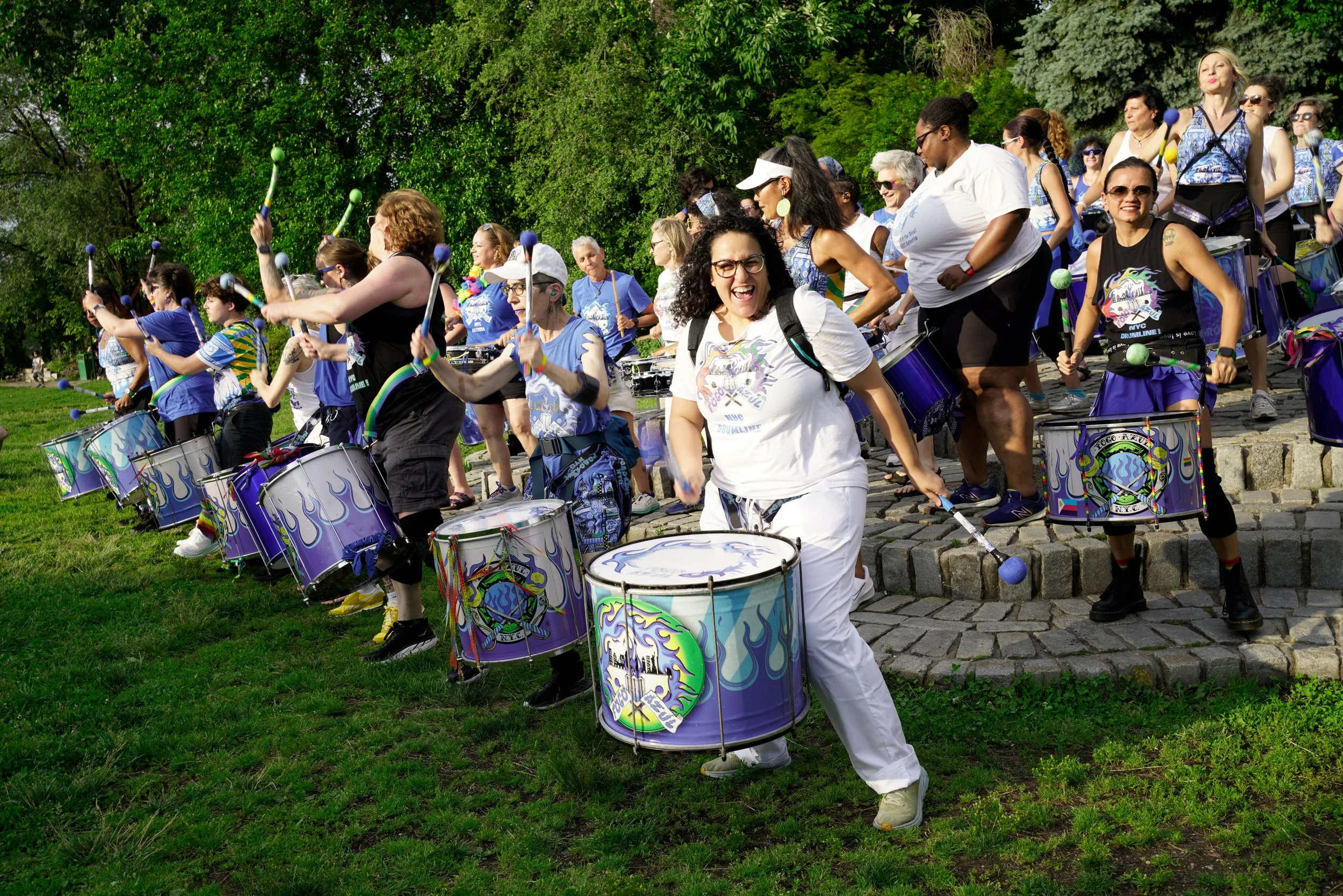 A group of diverse women playing drums outdoors in a park, participating in a drum circle event on a sunny day with green trees in the background.