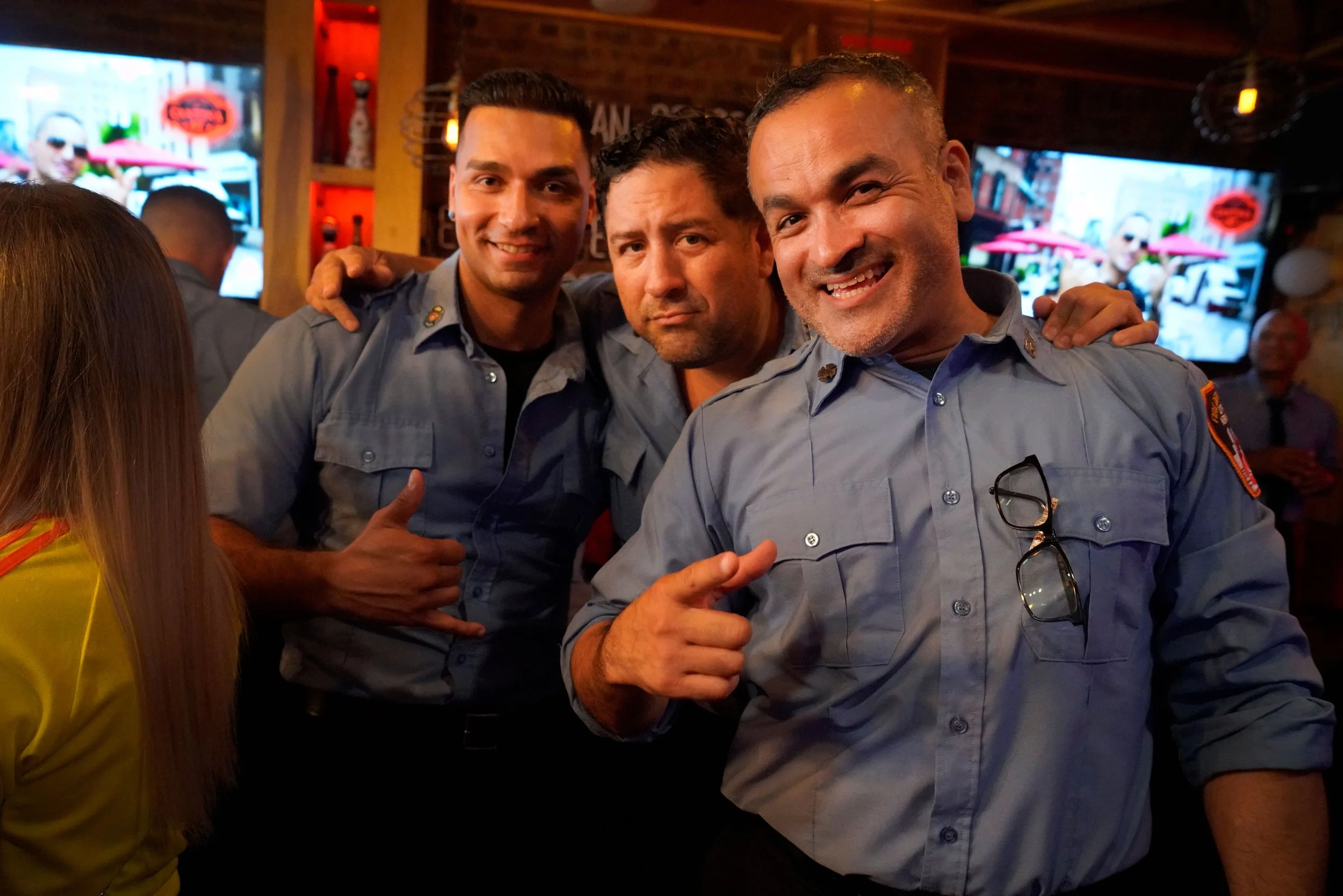 Three smiling police officers in uniform posing together in a bar or restaurant with a brick wall and TV screens in the background.