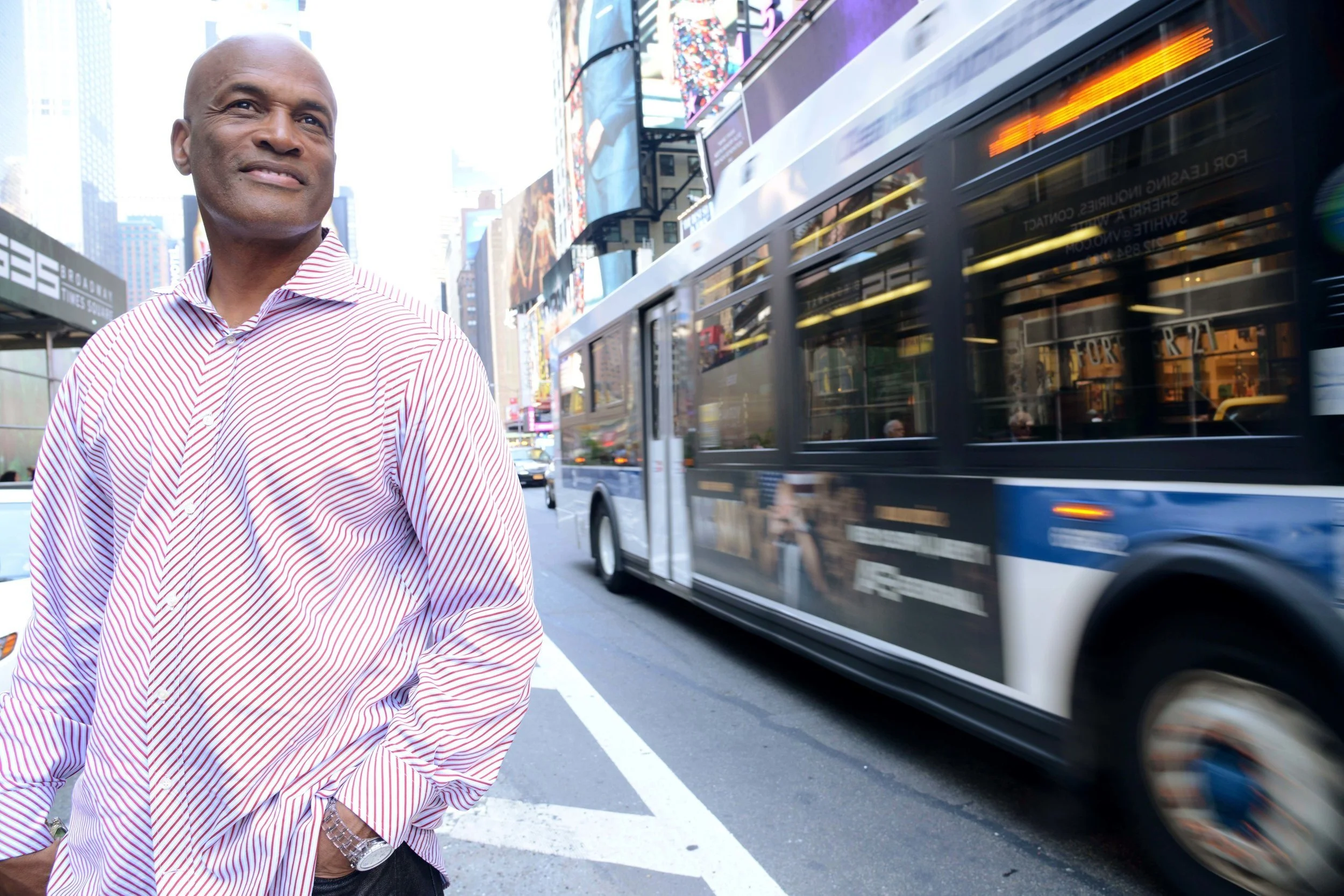 A man in a red and white striped shirt standing on a busy city street with a double-decker bus passing by