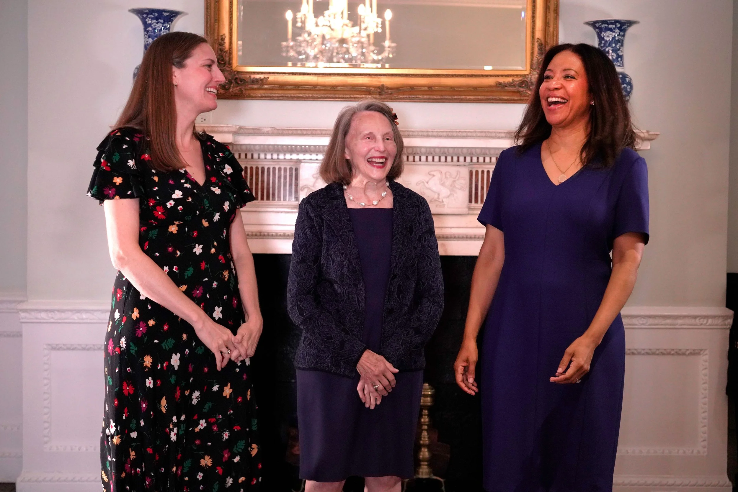 Four women standing and smiling in front of a fireplace with a mirror, chandeliers, and vases in the background.