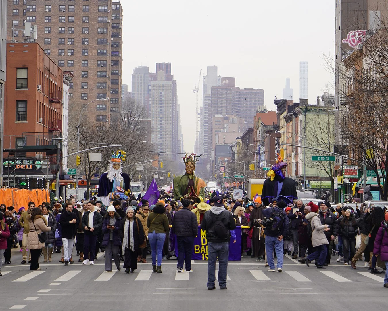 A street in a city filled with people participating in a parade or protest, with large puppets and banners, surrounded by tall buildings on a cloudy day.