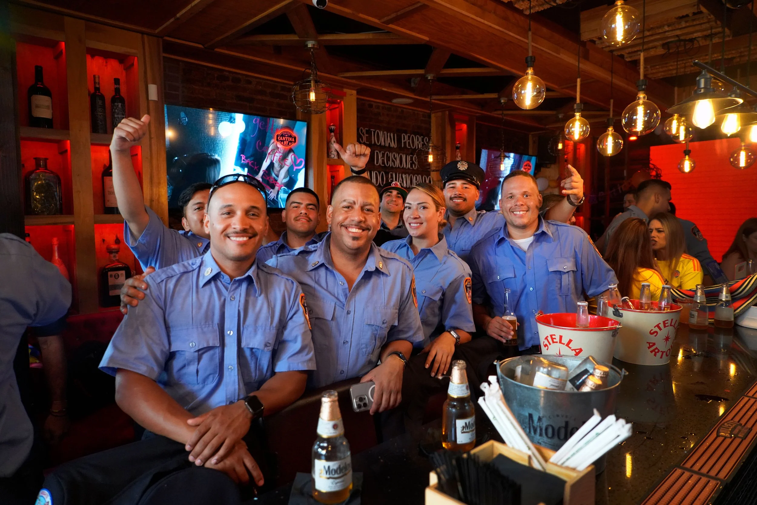 Group of smiling uniformed police officers inside a bar or restaurant, with bottles on shelves, flat screens, and warm industrial decor.