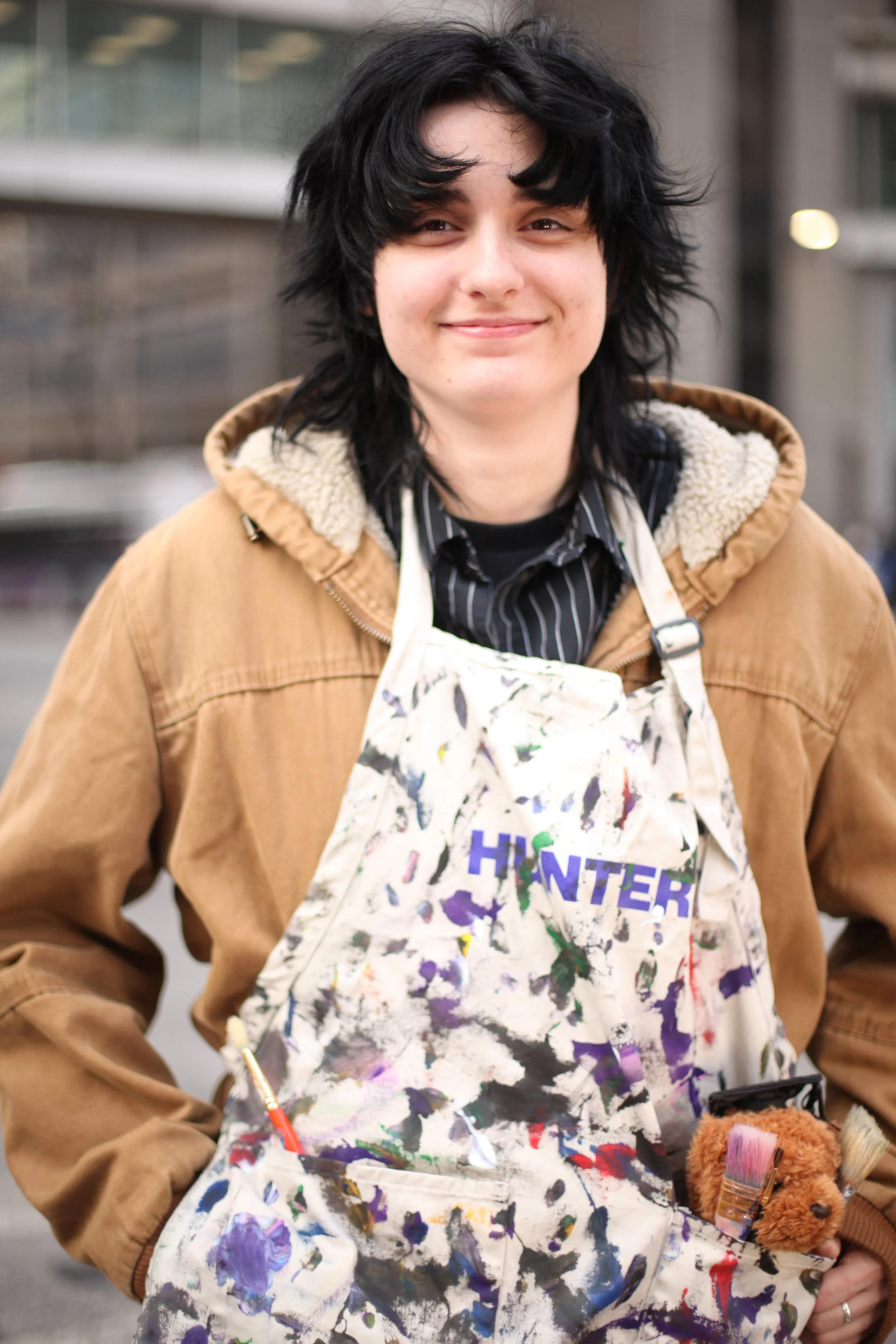 Young person with black curly hair wearing a tan jacket and colorful splattered apron, holding a paintbrush and a small stuffed animal in an outdoor setting.