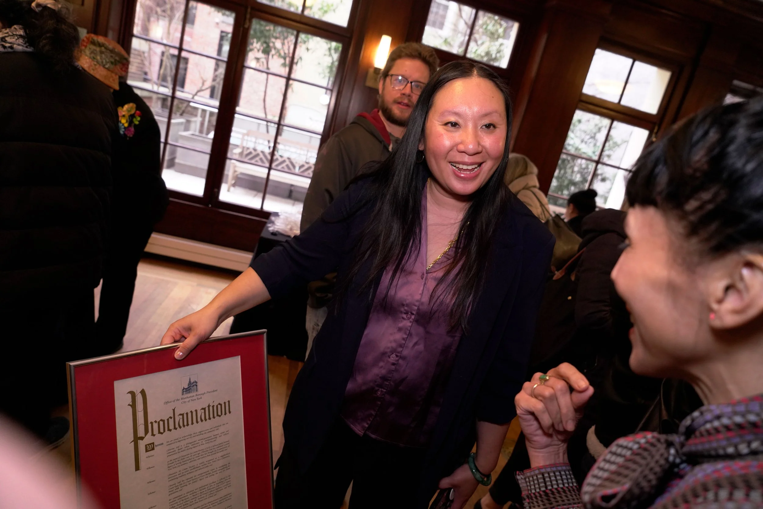 A woman with long black hair smiling and wearing a purple blouse, speaking with a person with dark hair and a plaid shirt. Behind her, a man with glasses and a beard is looking in her direction. The setting appears to be indoors with large windows an
