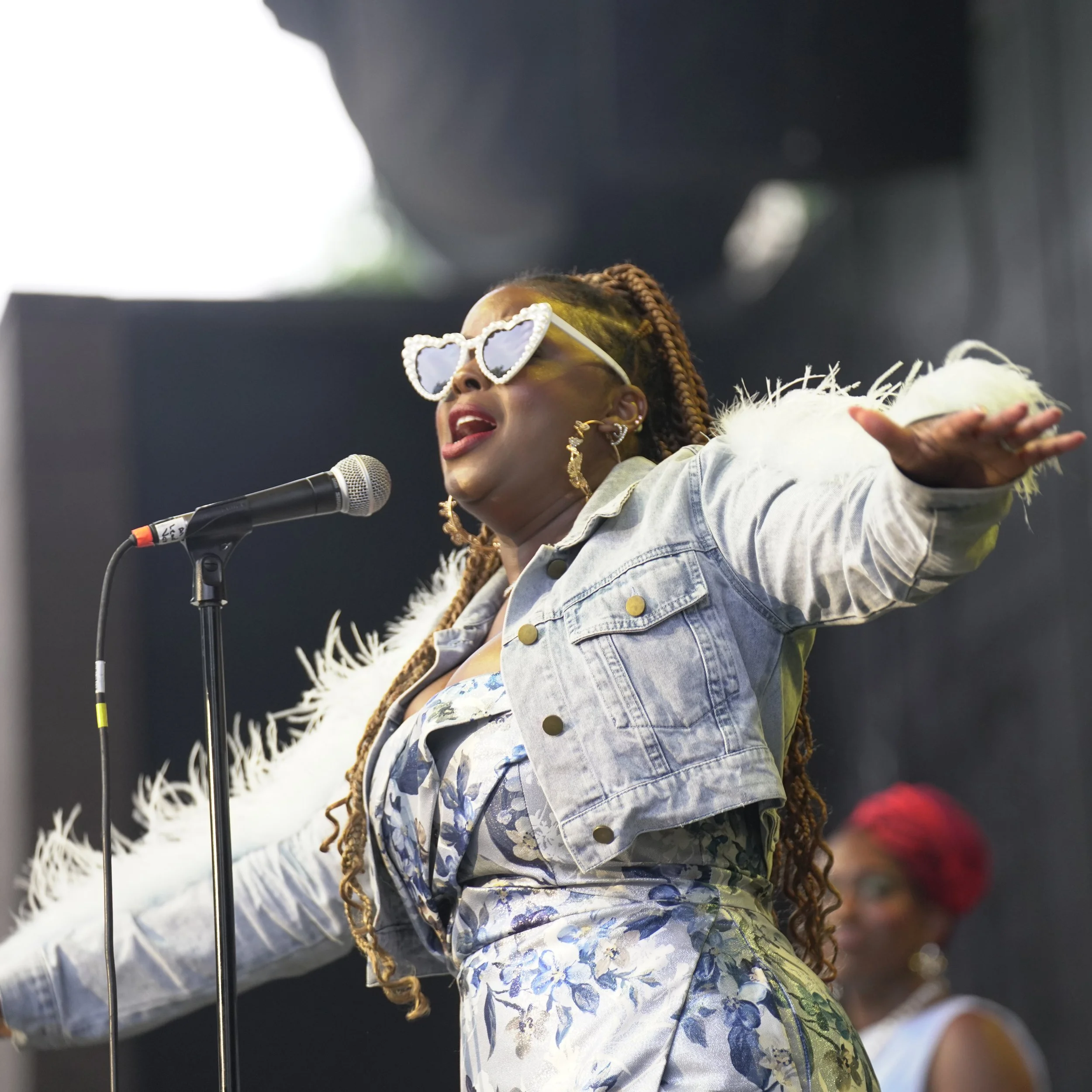 A woman with braided hair and heart-shaped sunglasses singing into a microphone, wearing a denim jacket with a patterned shirt and a fur-lined coat.