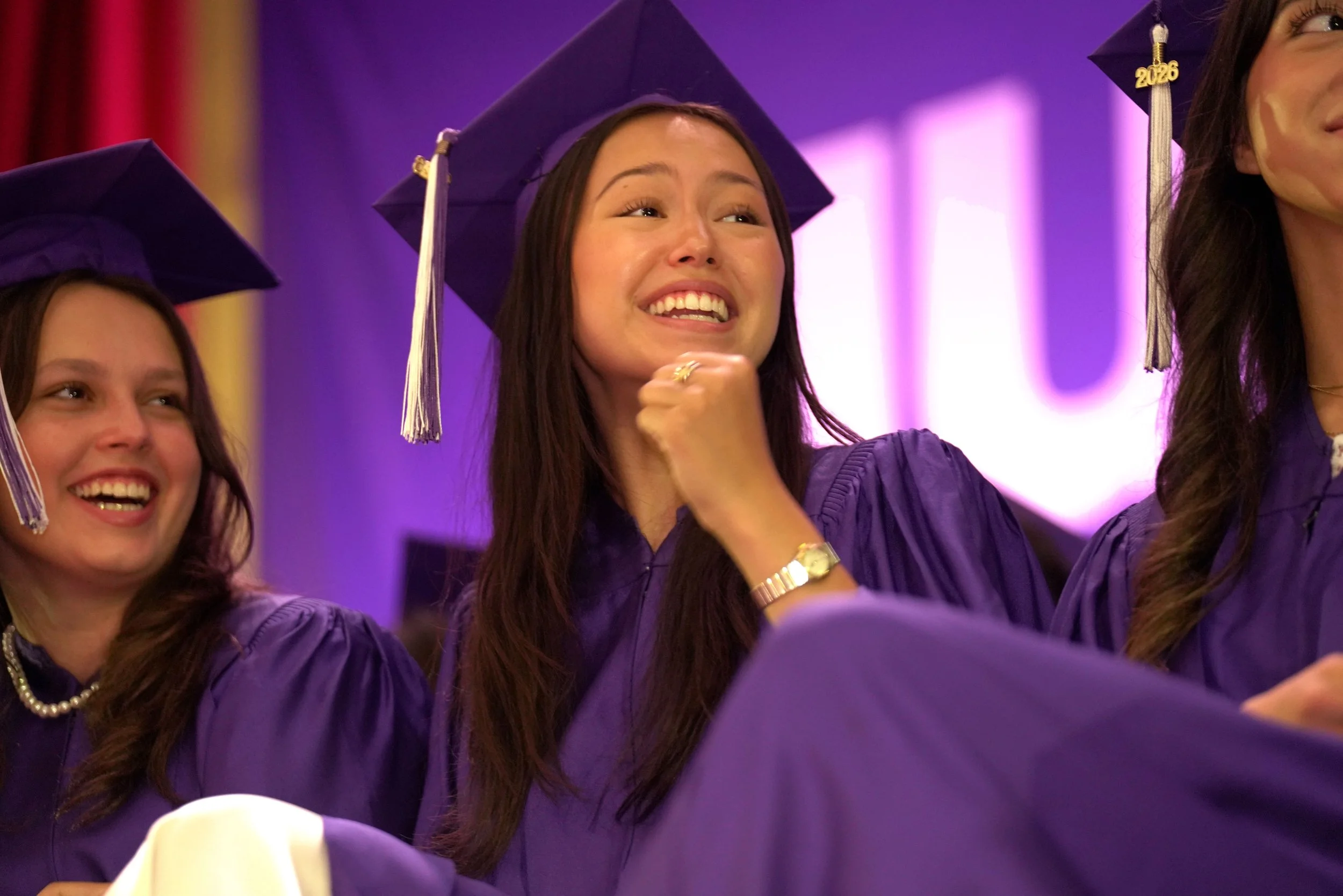 A group of female graduates wearing purple caps and gowns at a graduation ceremony, with one woman at the center appearing emotional and smiling.