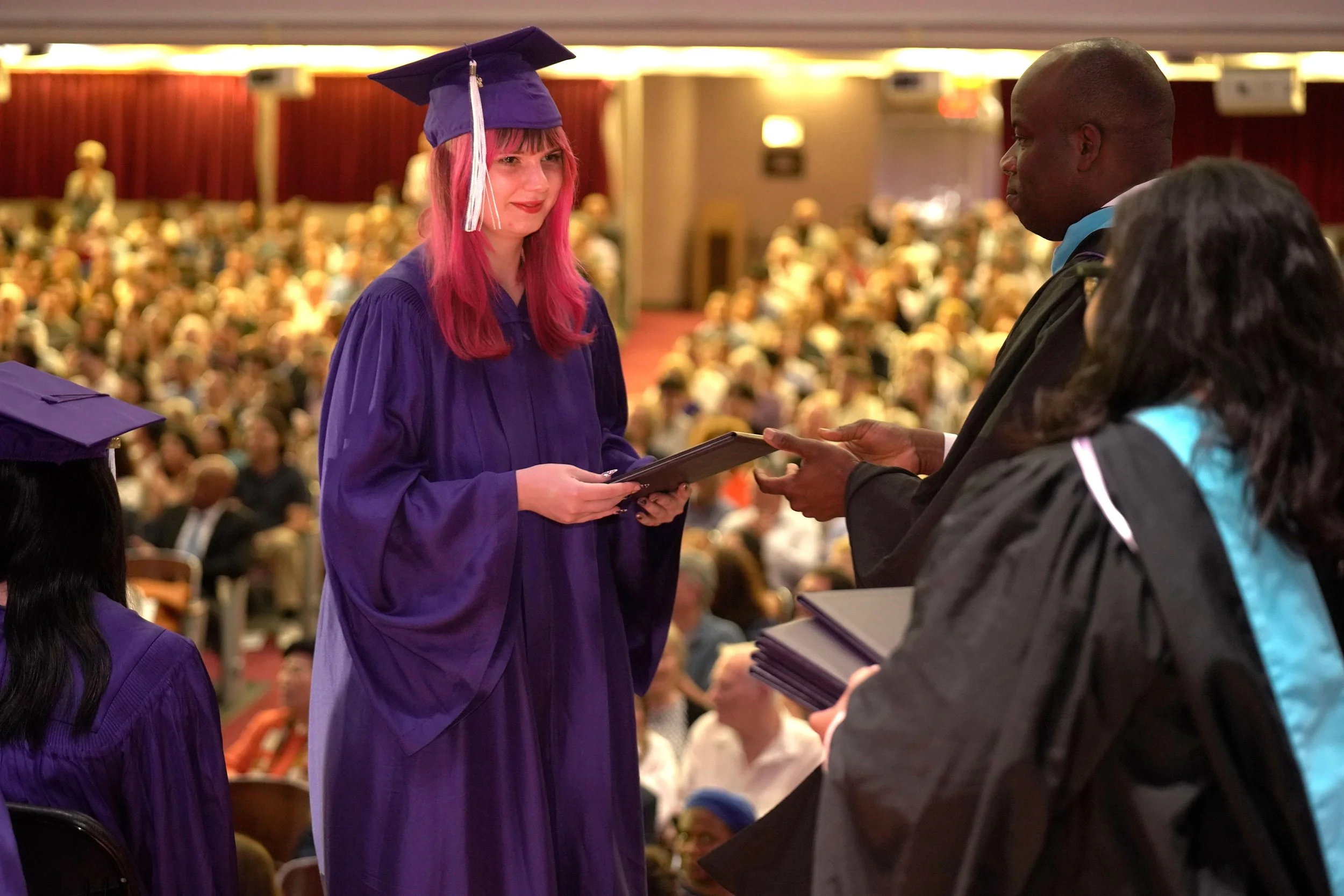 A young woman with pink hair receiving a diploma at a graduation ceremony, dressed in a purple gown and cap, with an audience in the background.