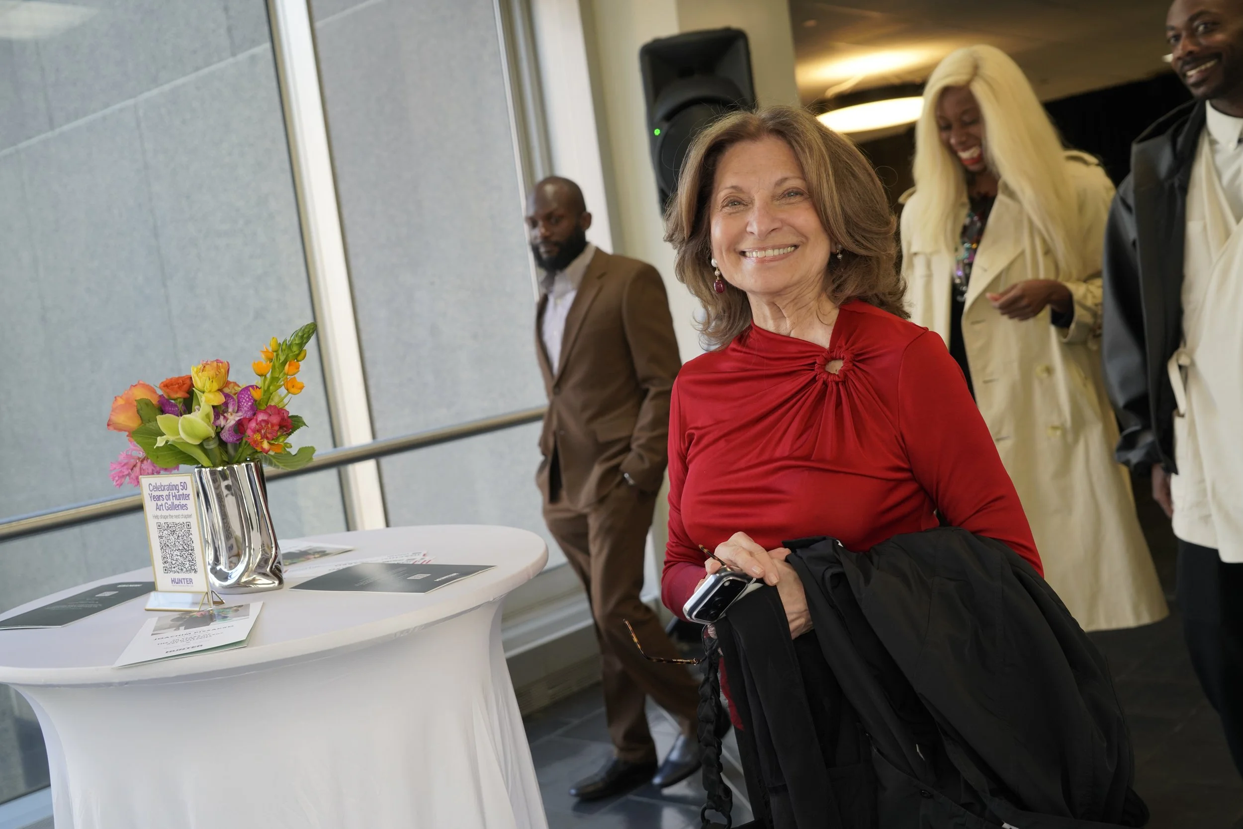 Smiling woman at a table with a bouquet of colorful flowers, in an indoor event space with other people in the background.