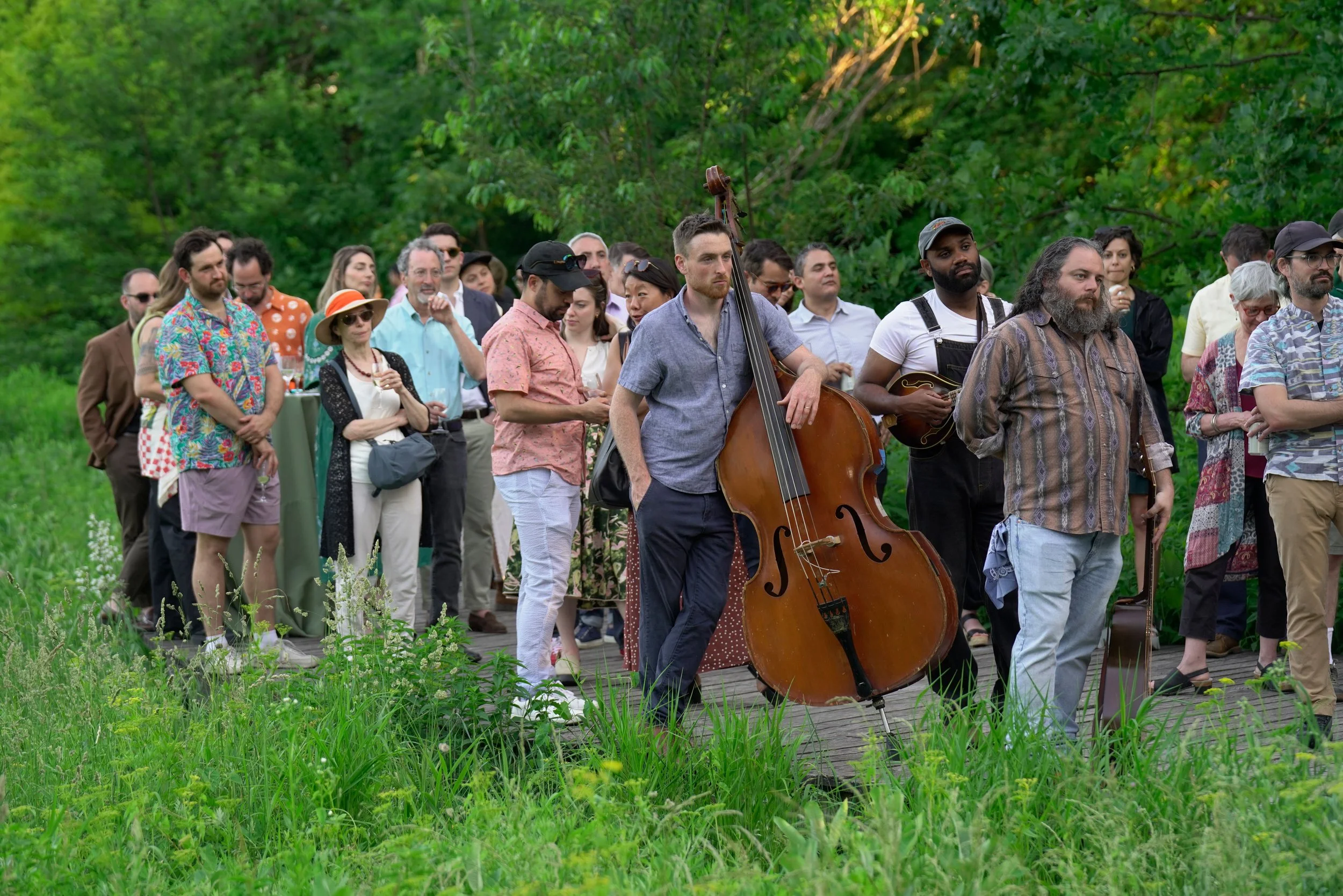 A group of people gathered outdoors on a wooden path, some holding musical instruments like a double bass and mandolin, surrounded by green trees and grass.