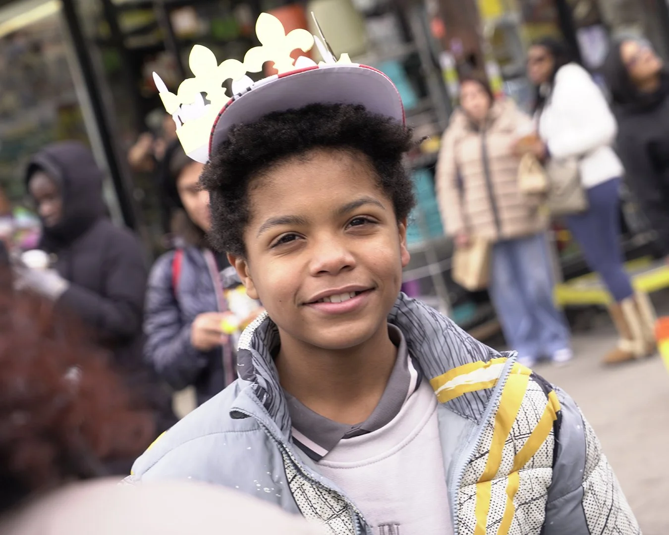 A young boy with short curly hair smiling, wearing a birthday crown, standing on a sidewalk with people in the background.