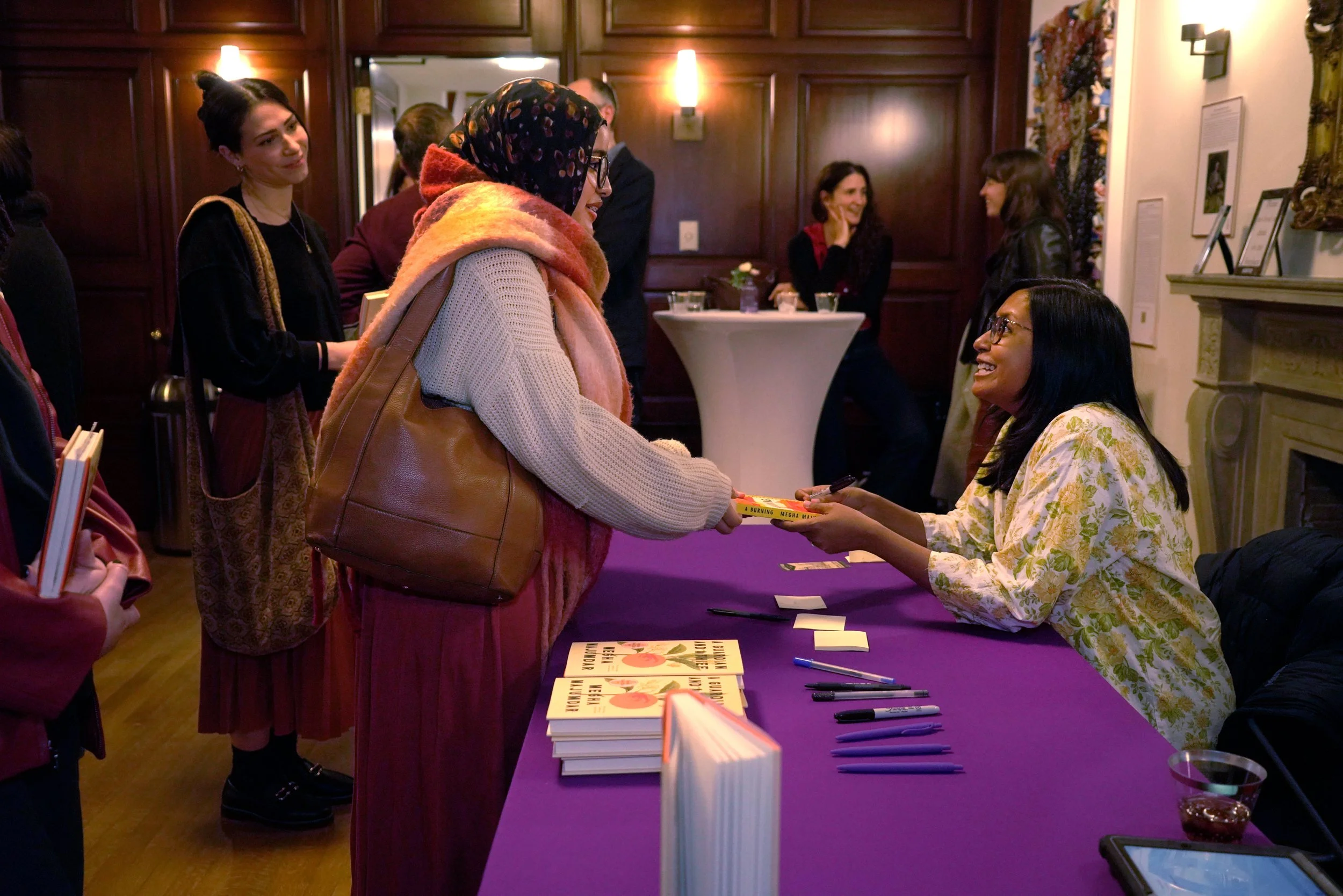 A woman in a colorful scarf and cream sweater is receiving a book from a woman seated at a table covered with books and writing materials in a room with wooden wall paneling. Other people are interacting and talking in the background.
