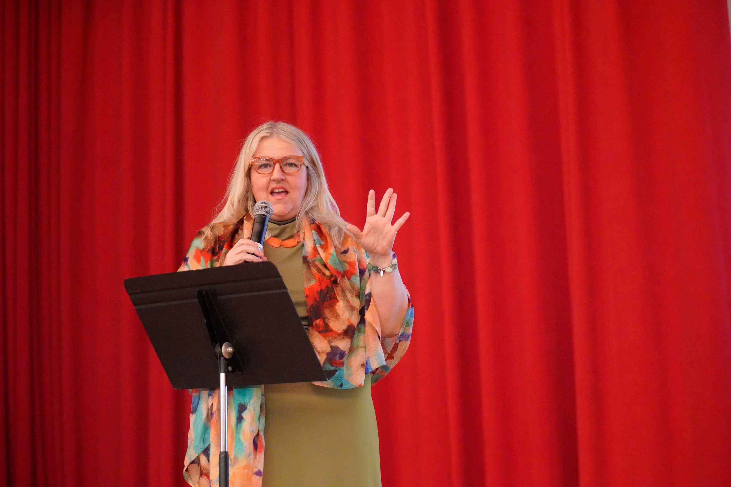 A woman with blonde hair, wearing glasses, a colorful floral jacket, and a green dress, speaking into a microphone on stage with a red curtain background.