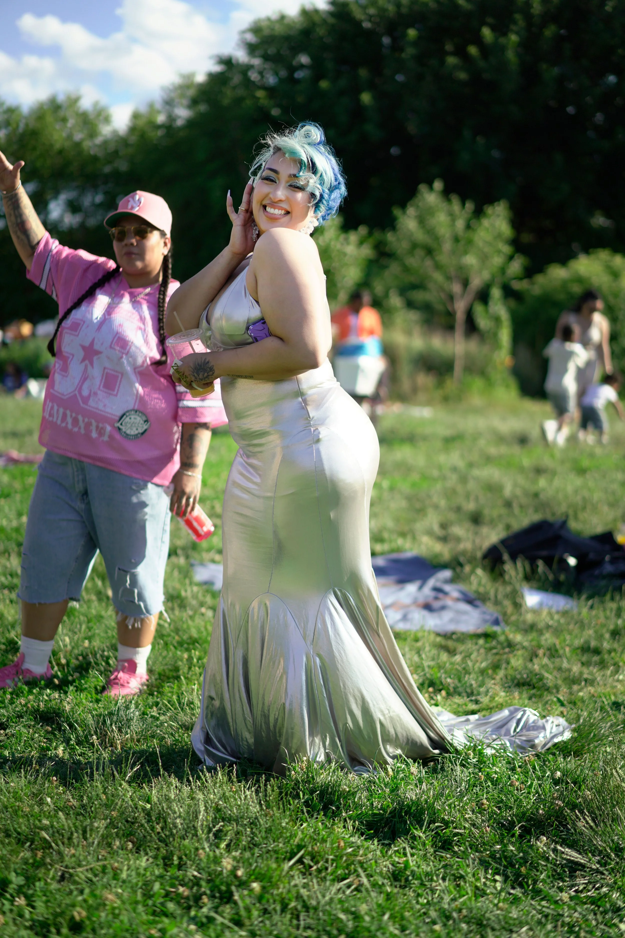 A woman with blue and green hair smiling and posing outdoors on a sunny day, wearing a shiny silver dress. Behind her, a person with sunglasses, braids, and a pink jersey poses with one arm raised. There are other people in the background in a park s