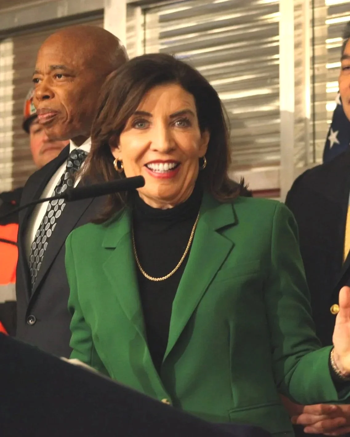 A woman with dark brown hair smiling and speaking at a microphone, dressed in a green blazer and black top, standing in front of a metal background with other people around.