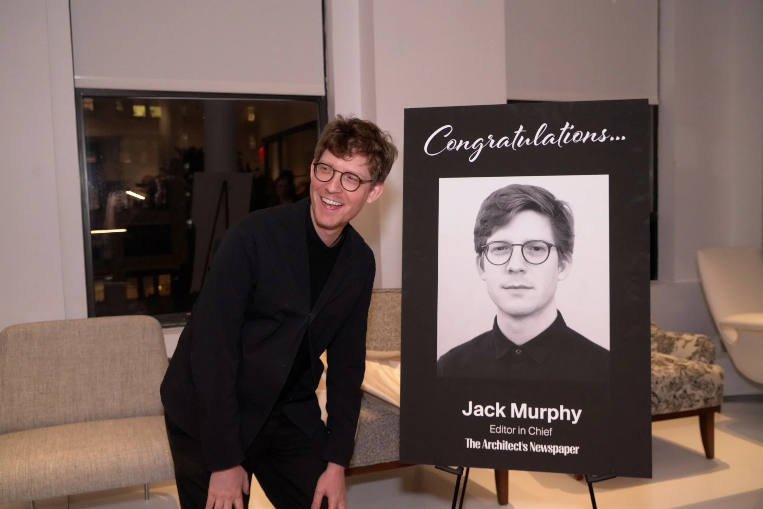 A man with glasses and curly hair smiling and leaning forward at an event, standing next to a black and white poster with a portrait of a young man wearing glasses. The poster features the text 'Congratulations...' and the name Jack Murphy, Editor in