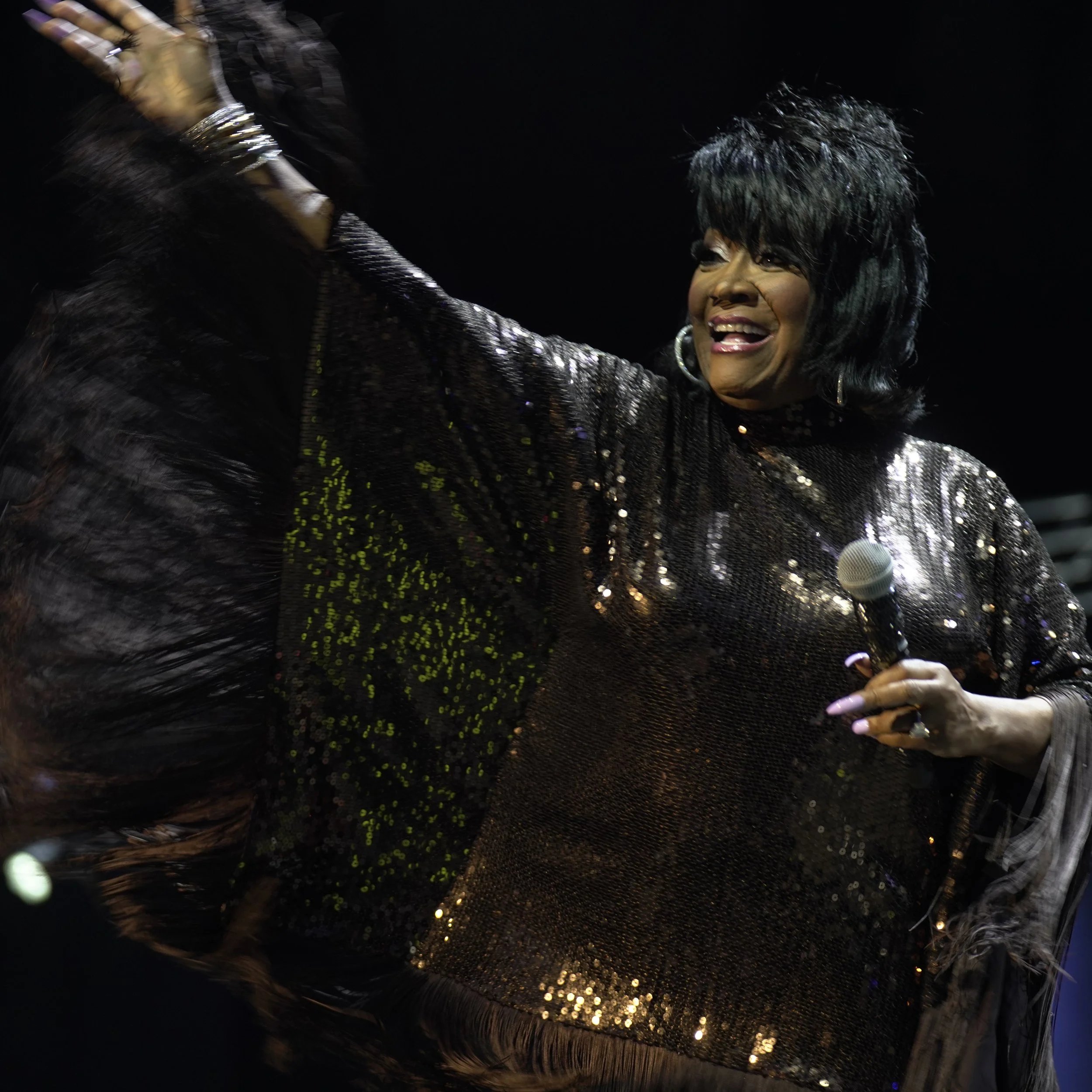 A joyful black woman with short curly hair and earrings, wearing a shiny black sequined dress, smiling and waving with her right hand, with a microphone visible in the lower right corner.