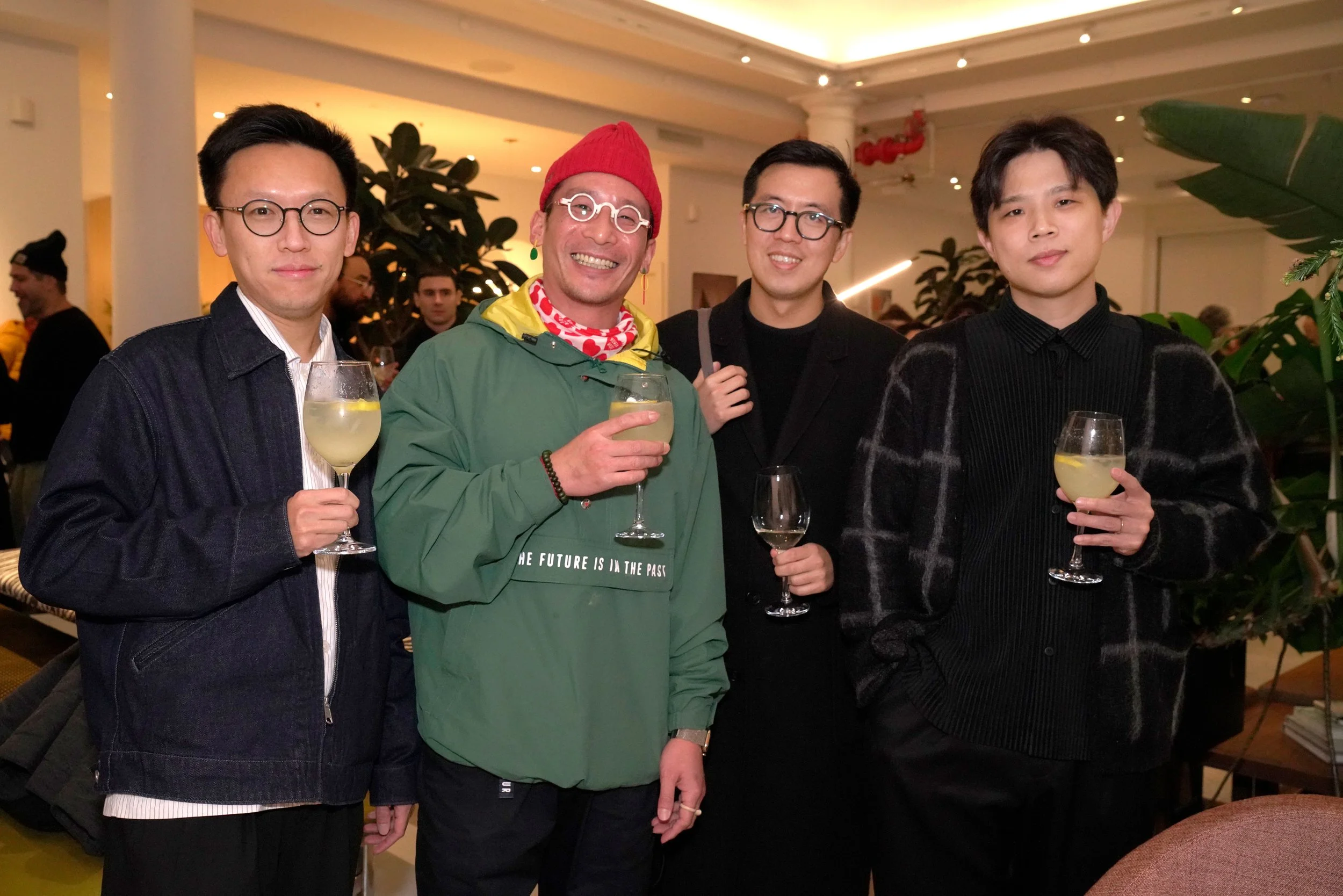 Four men standing together indoors at a social event, holding glasses of white wine, smiling, with plants and other people in the background.