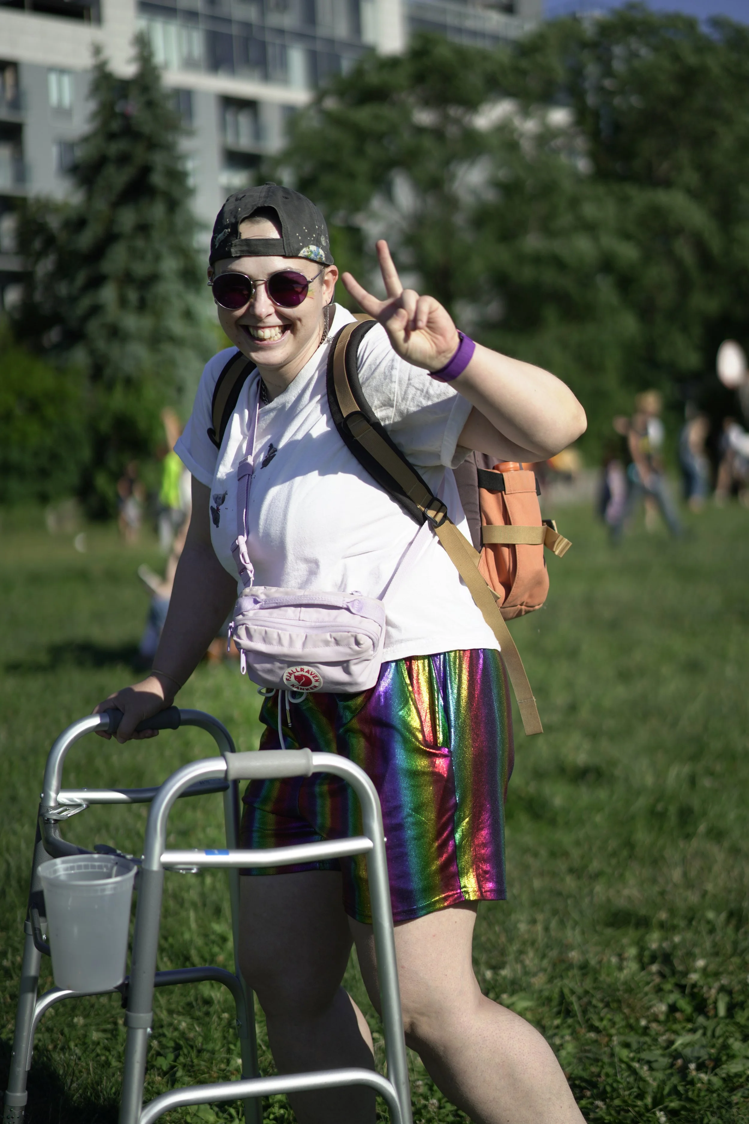 A smiling person with short hair wearing sunglasses, a baseball cap, a white T-shirt, rainbow-colored rainbow shiny skirt, and a purple wristband, using a walker outdoors on a grassy area with trees and buildings in the background, making a peace sig