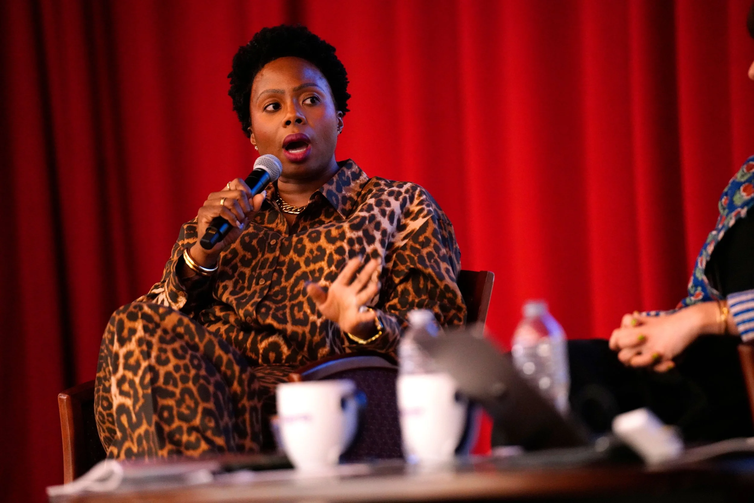 A woman wearing leopard print clothing speaking into a microphone during a panel discussion with a red curtain background.