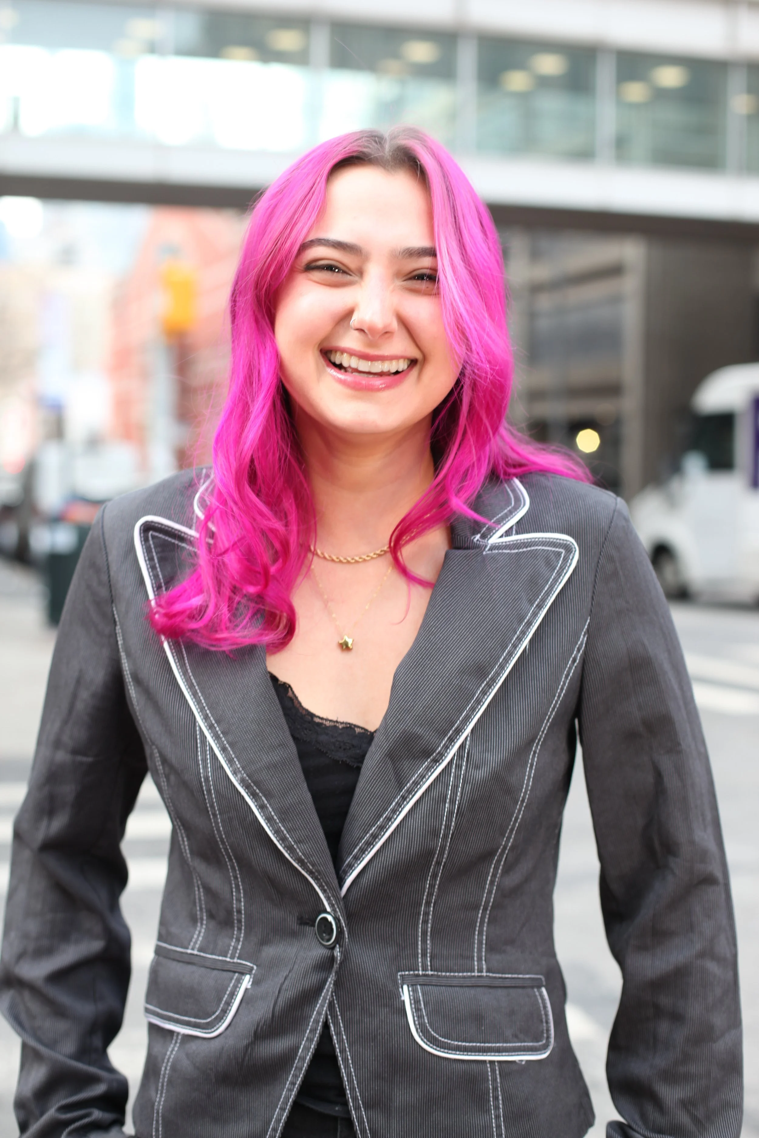 A woman with long, wavy pink hair smiling outdoors near a city street.