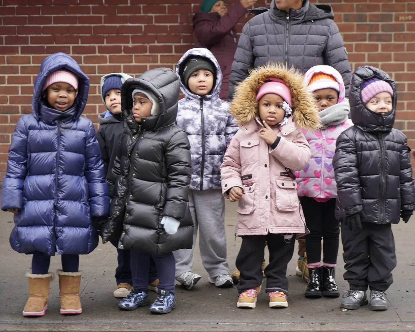 Group of young children standing outdoors in cold weather, wearing winter coats, hats, and boots, with a brick wall in the background.