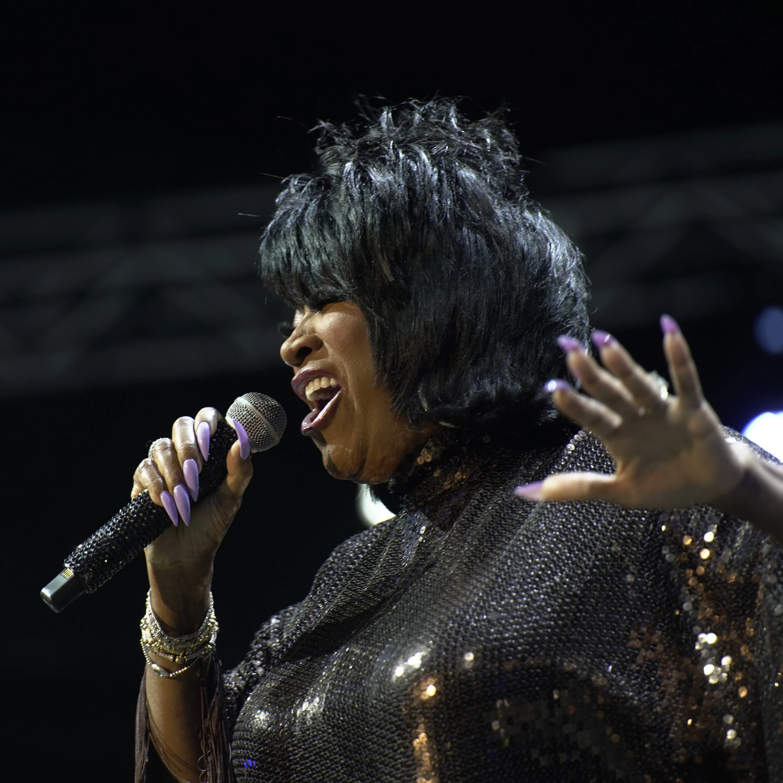 A woman singing into a microphone, with her hand raised, wearing a glittering black outfit and jewelry, against a dark background.
