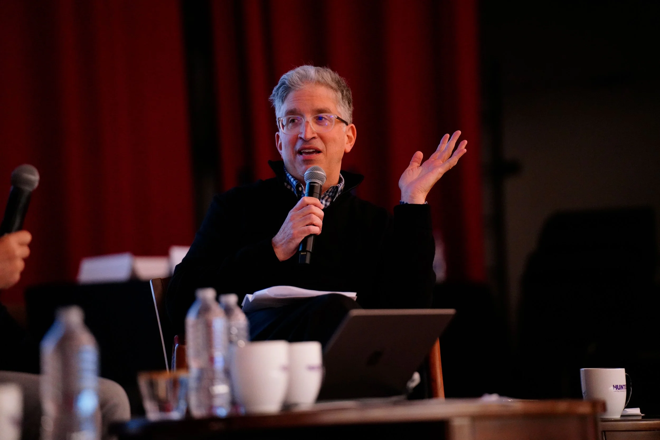 A man holding a microphone is speaking or presenting at a conference or panel, seated at a table with water bottles and papers, with a red curtain in the background.