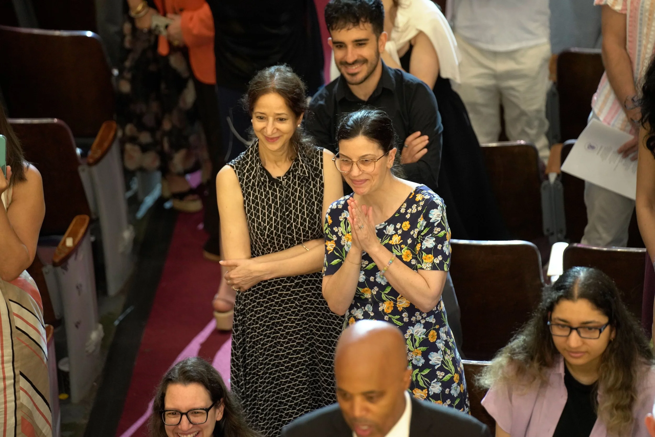A group of people at a crowded indoor event, with three women in the center showing expressions of happiness and prayer, all framed by other attendees around them.