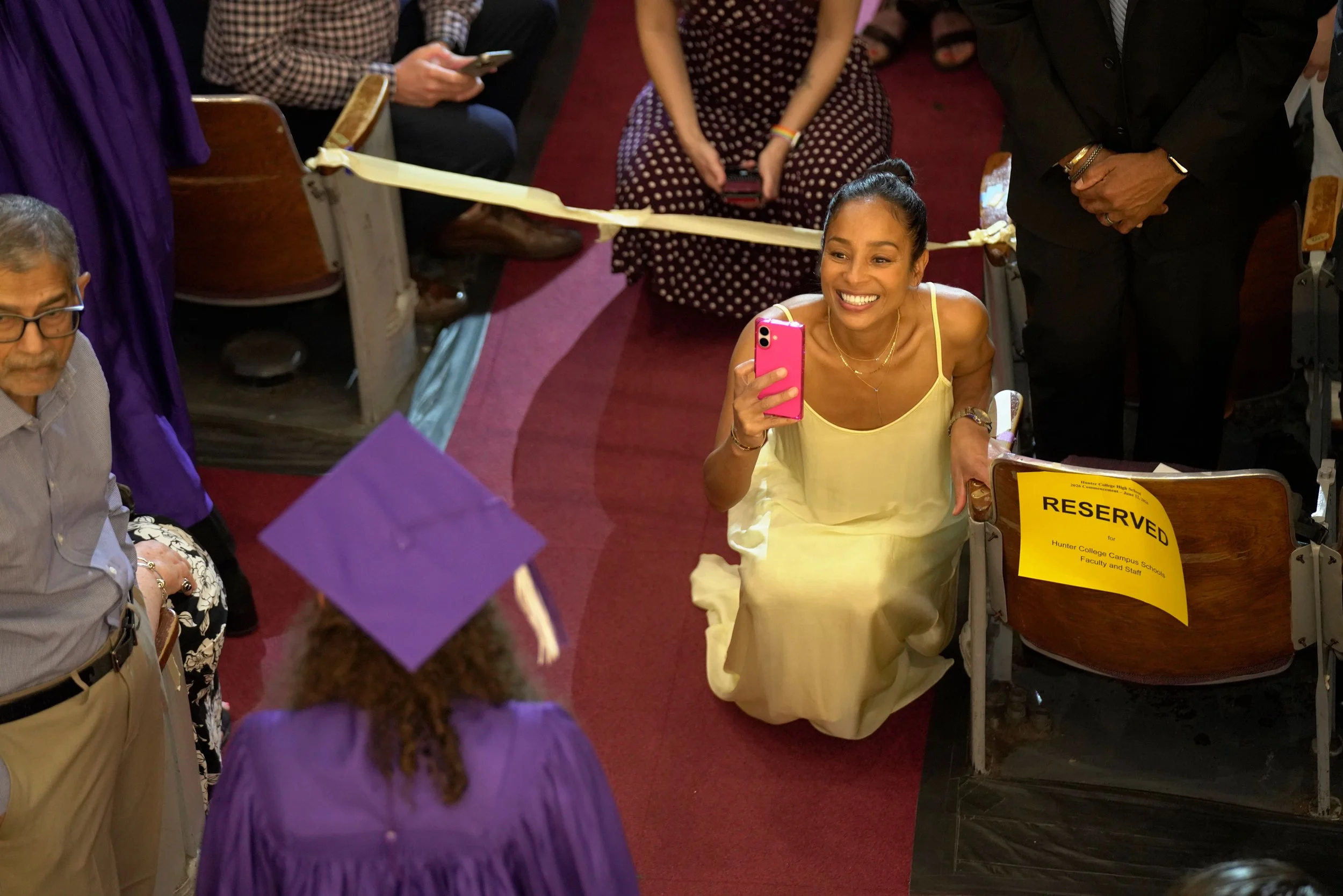 A group of people gathered in a conference or event setting, with a woman in a yellow dress smiling and taking a selfie with her phone while others are engaged in conversation and seated around her. There is a reserved sign on a table in the foregrou