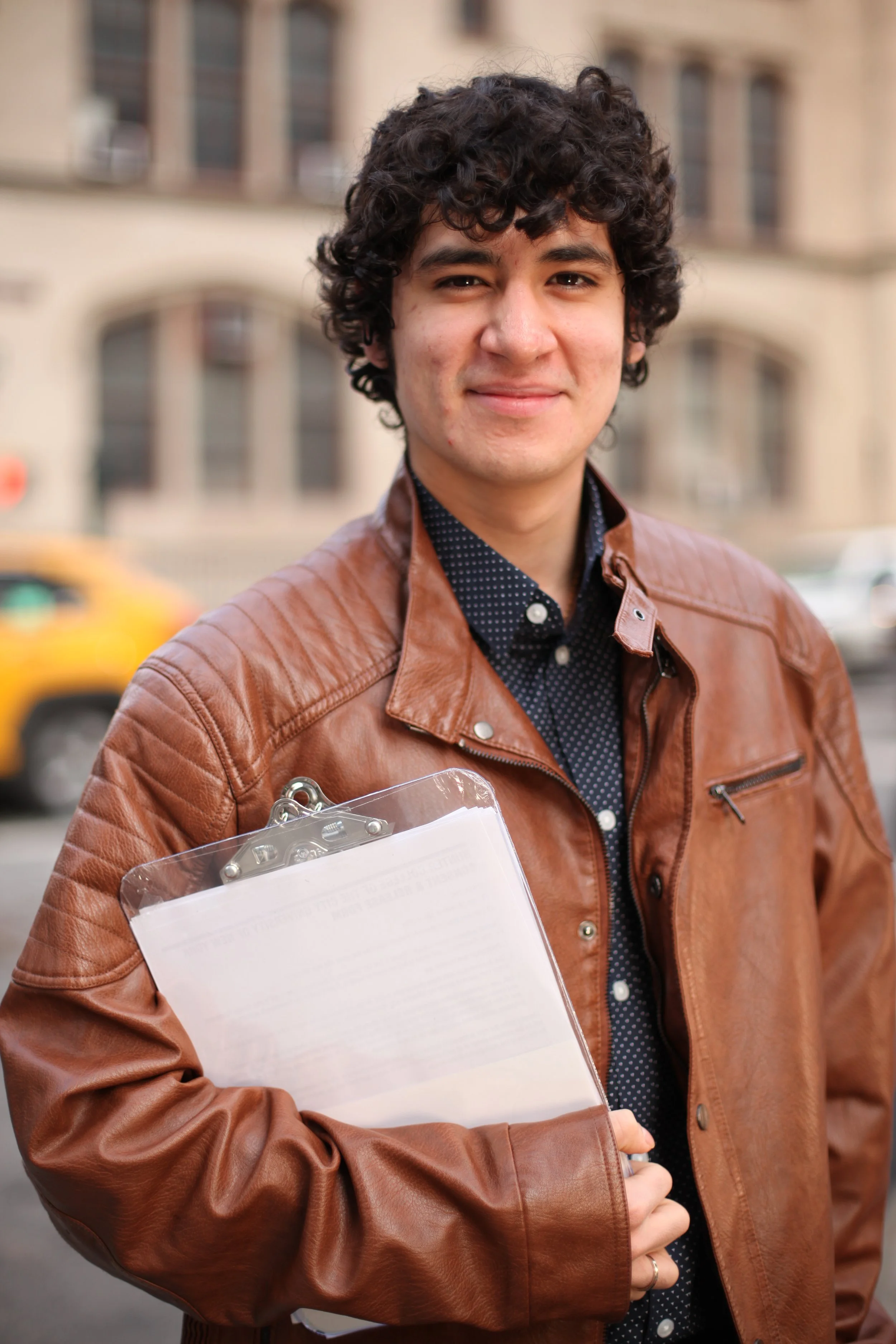 A young man with curly dark hair, wearing a brown leather jacket and a dark blue polka-dot shirt, holding a clipboard, standing outdoors with a blurred building and yellow taxi in the background.