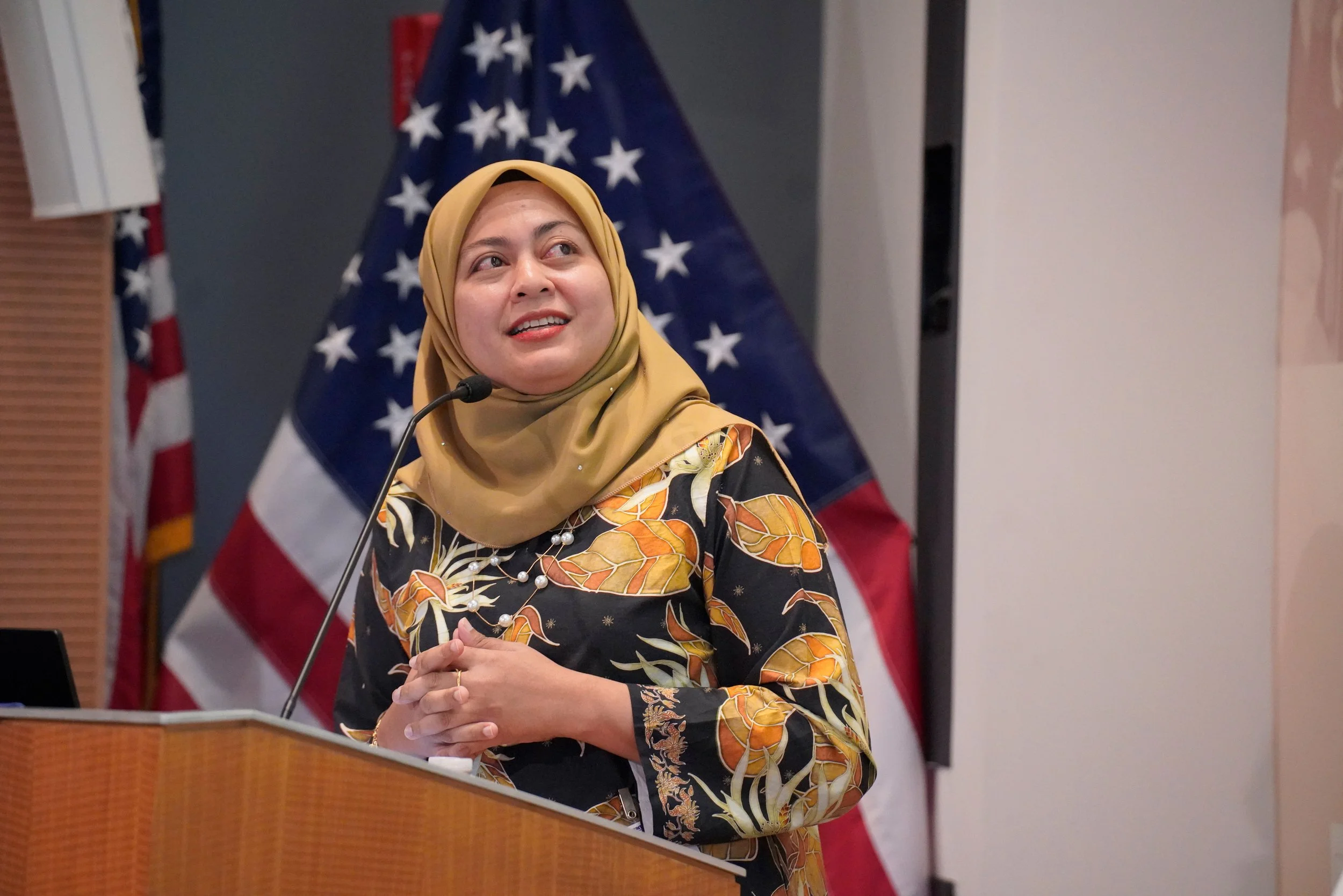 A woman wearing a tan headscarf and a black dress with a yellow and orange leaf pattern, standing at a podium with her hands clasped. Behind her are multiple American flags.