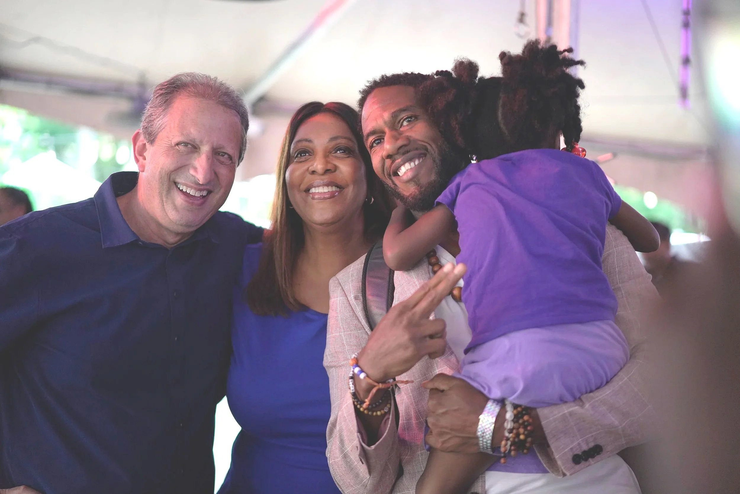 Three people smiling and embracing each other at an event, with a young boy being held by one of the women.